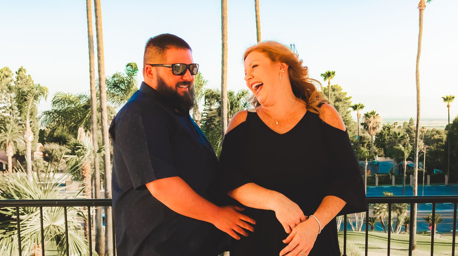 A happy couple laughing together outdoors on a sunny day with palm trees in the background.