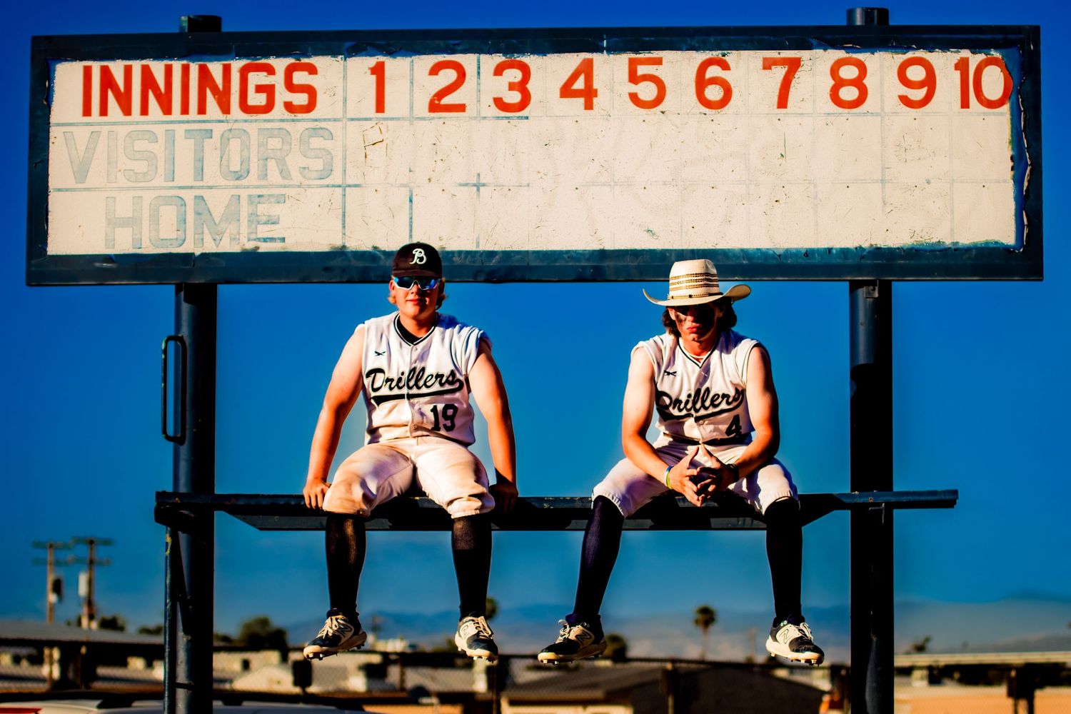 Two baseball players sit on the dugout bench beneath a scoreboard during a sunny day at the ballpark.