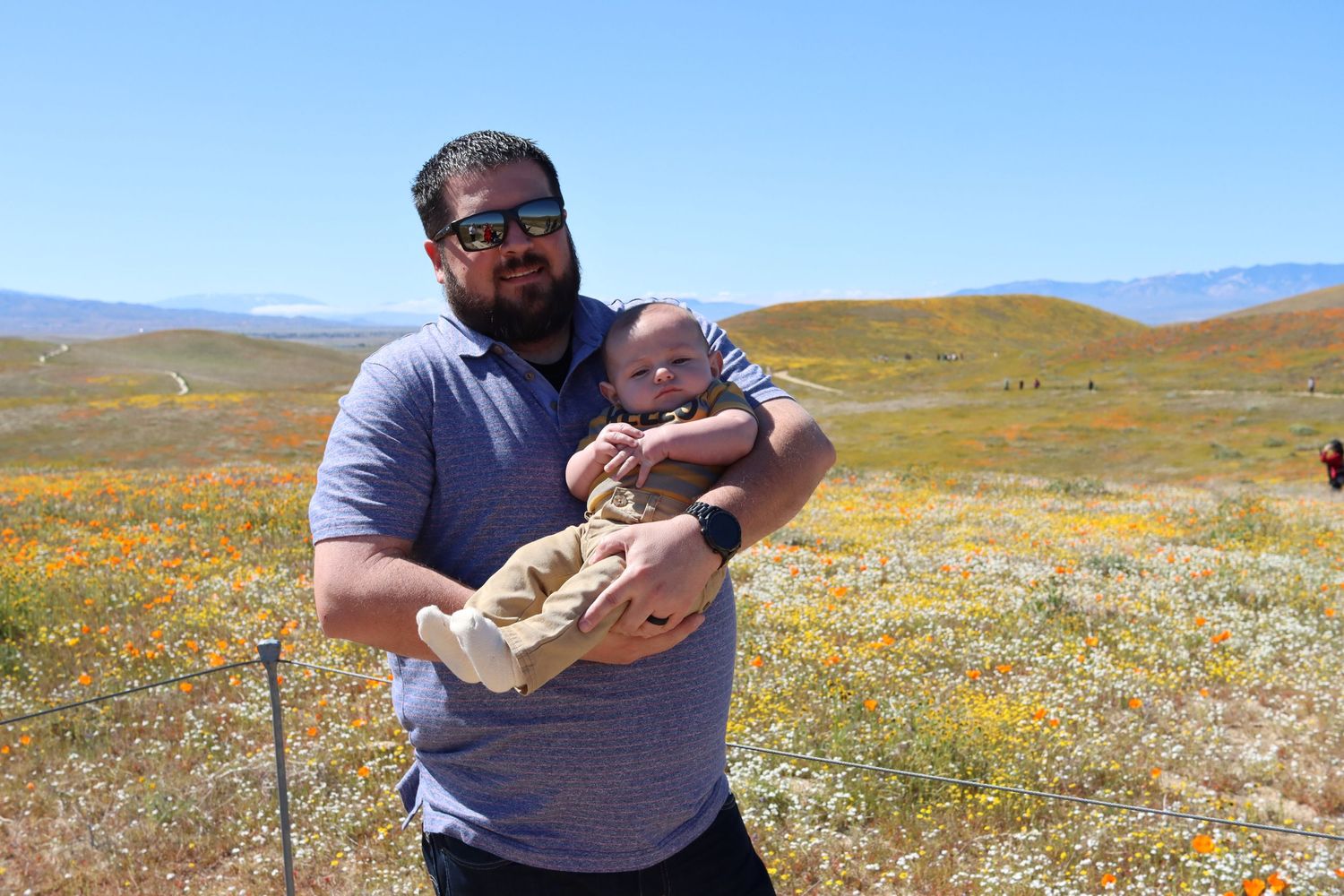 Two people standing in a field of orange wildflowers with mountains visible in the background on a sunny day.