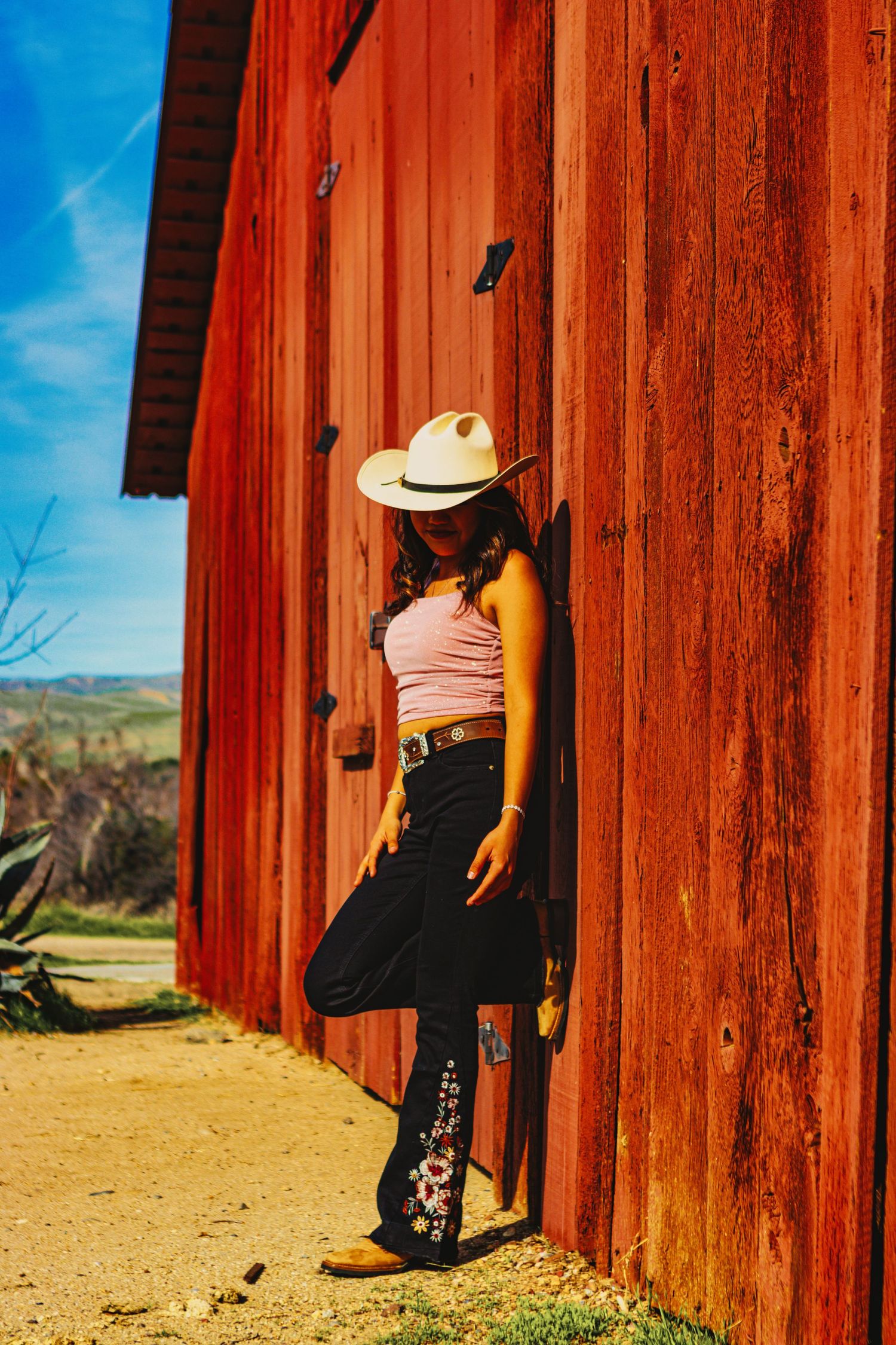 Person in cowboy hat and jeans leaning against vibrant red barn wall in sunlight.