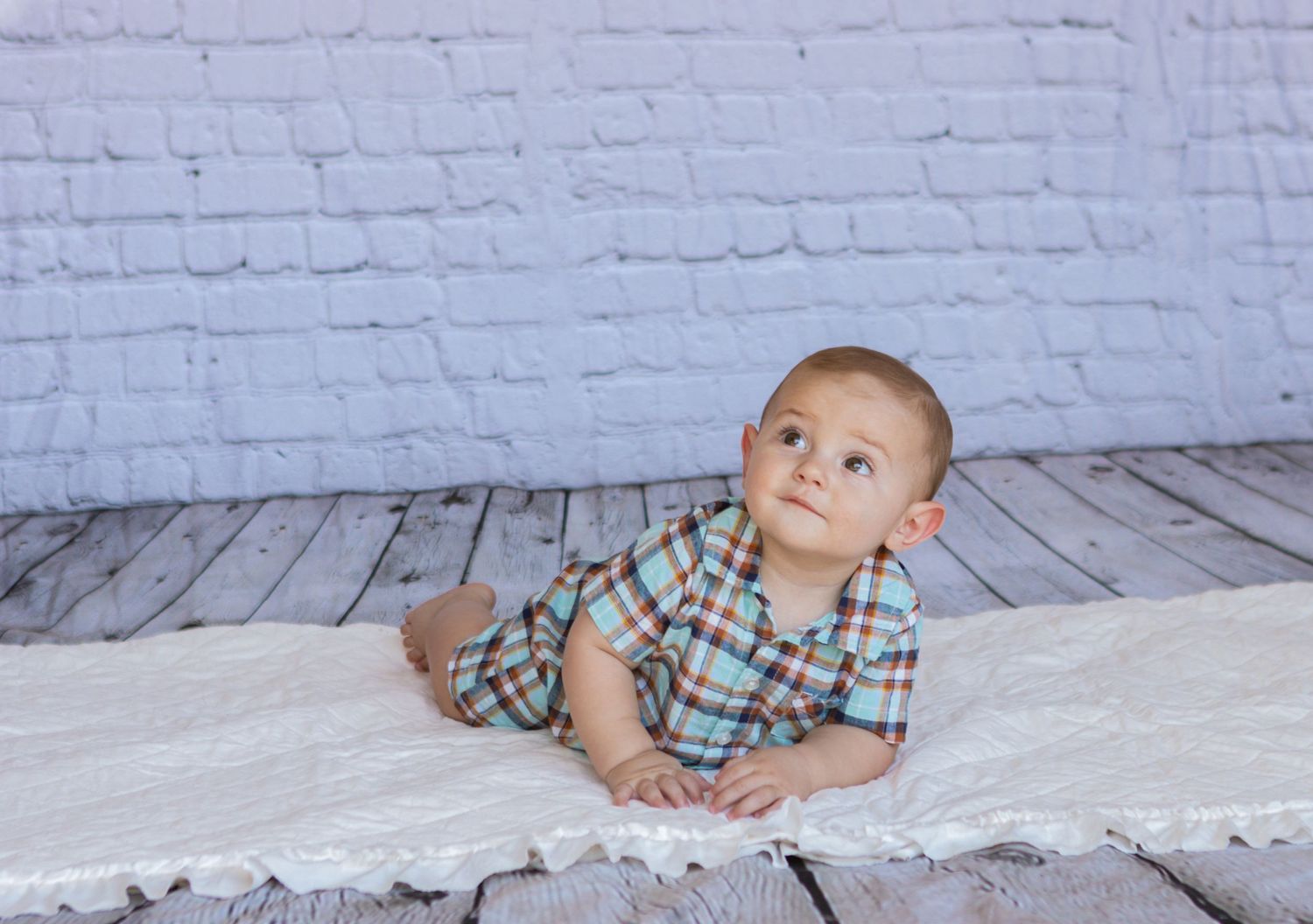 Baby in checkered outfit poses on white wooden floor against brick wall.