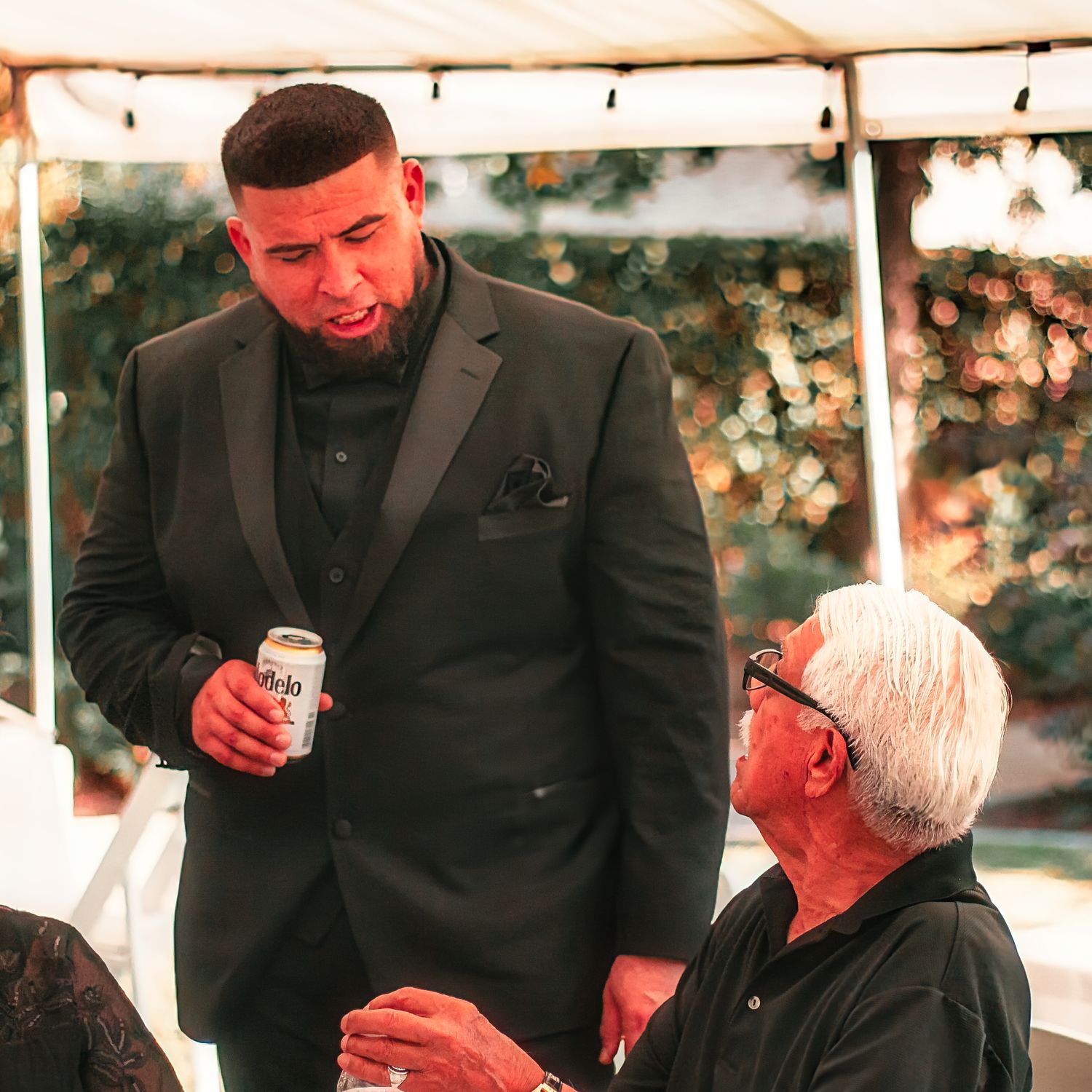 Two people in formal attire share a conversation under a tent canopy during a daytime outdoor event.