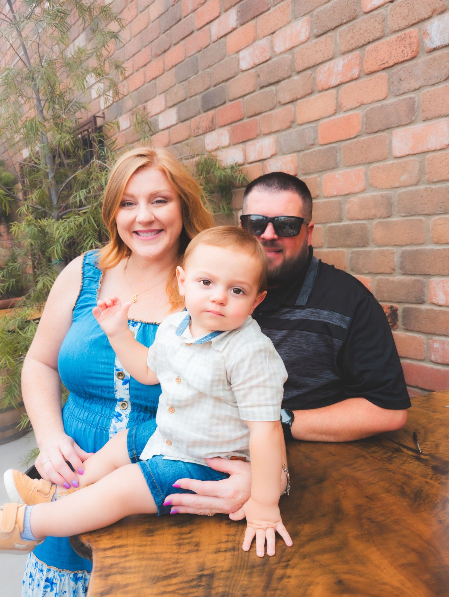 A family group sits together on a wooden bench against a brick wall on a sunny day.