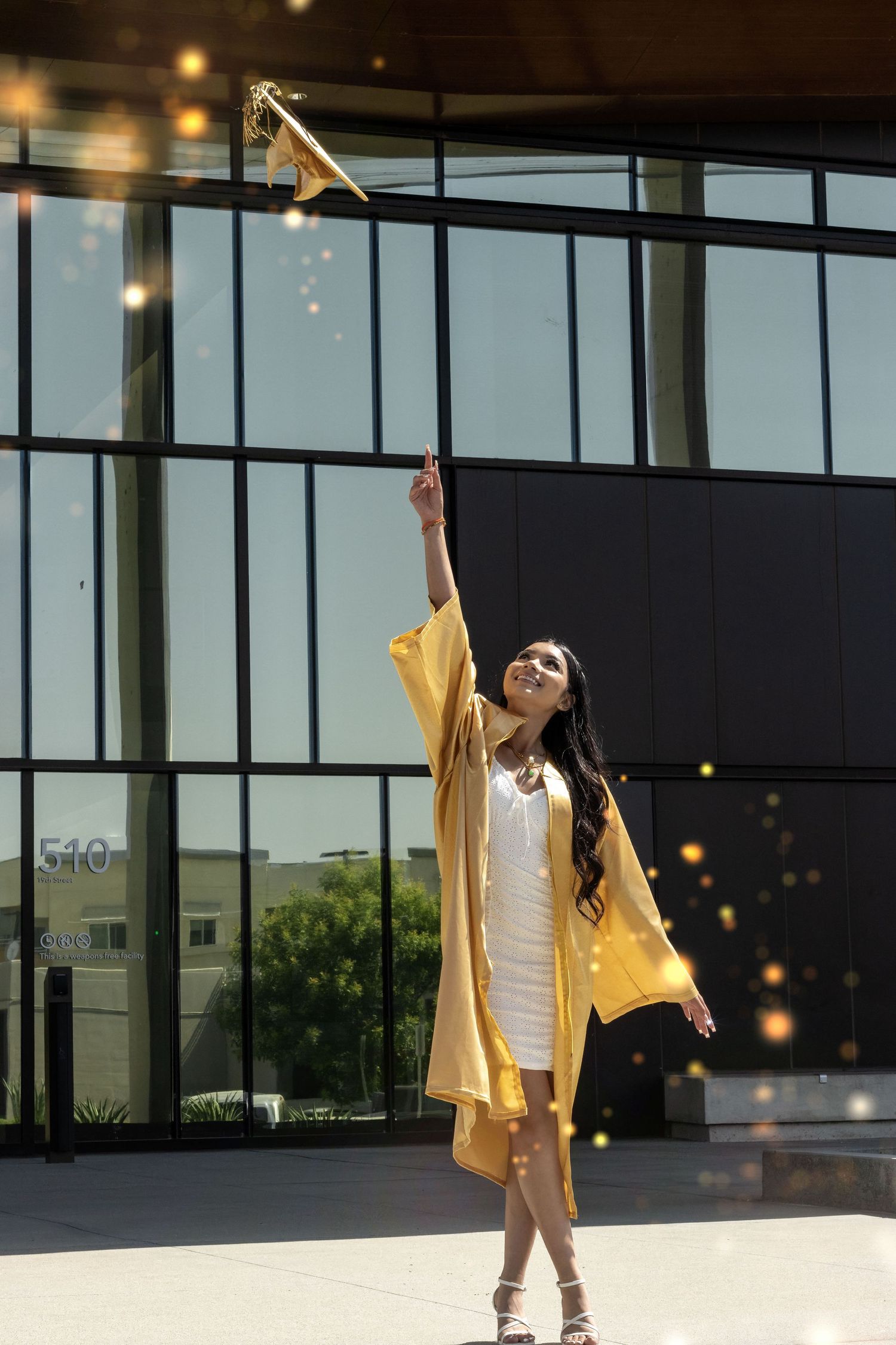 Graduate in yellow cap and gown holds diploma up high while celebrating outside modern glass building.