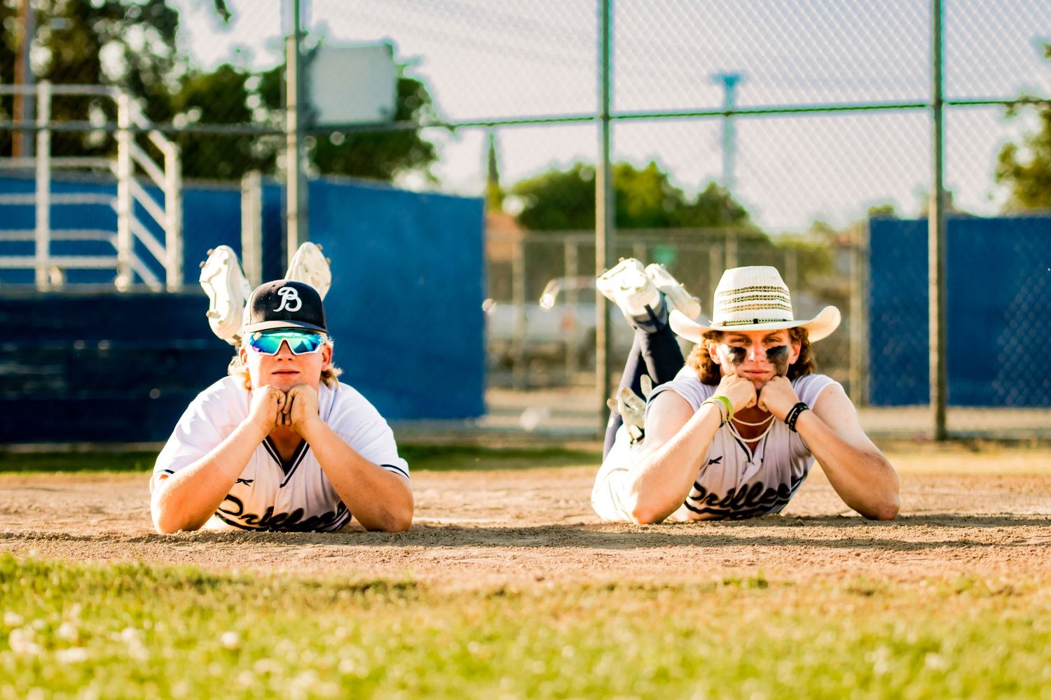 Two youth baseball players lying on their stomachs in the dirt on a baseball field with gloved hands under their chins.
