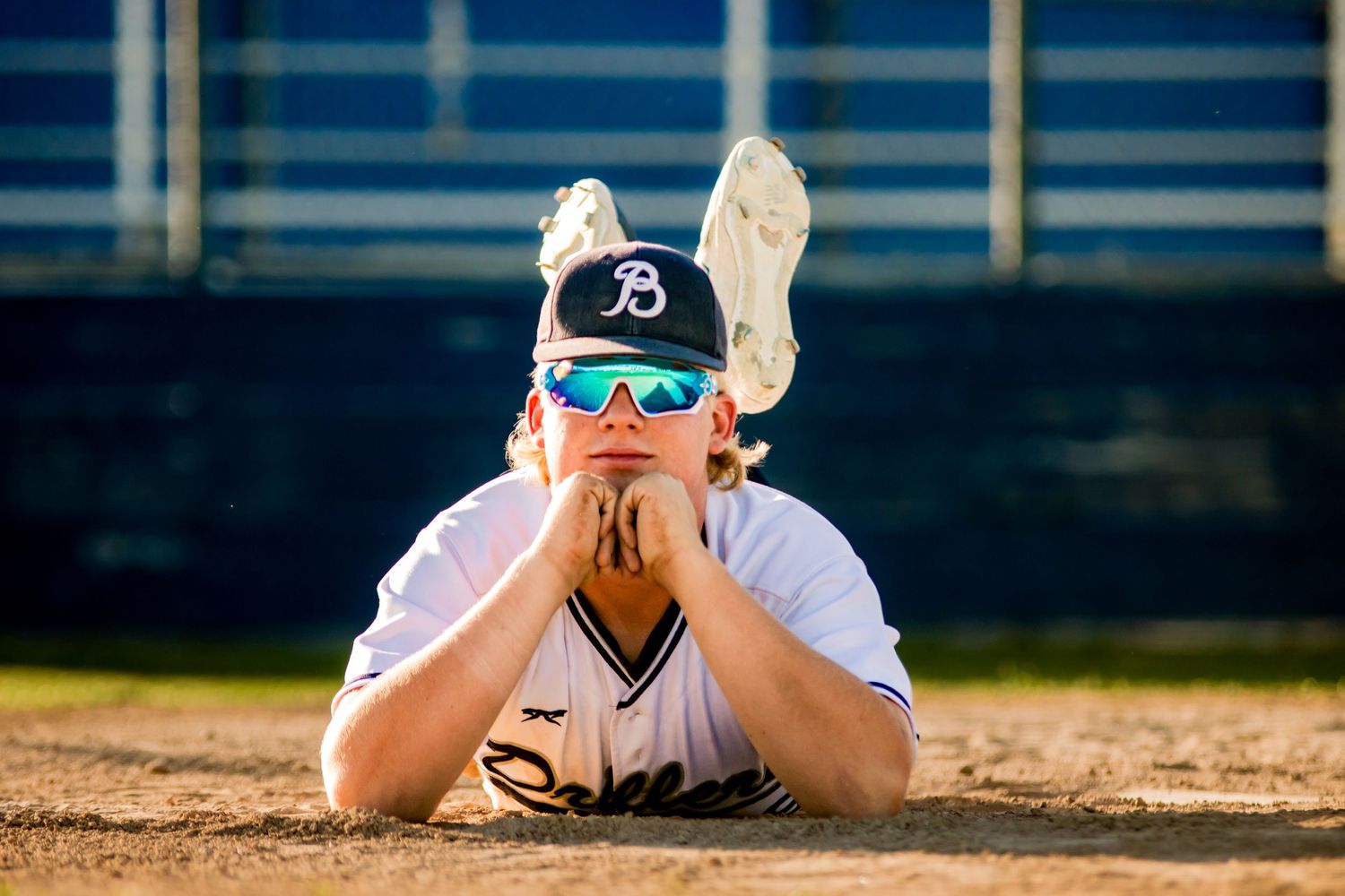Baseball player in white jersey and sunglasses lying on dirt field with a dugout in the background.