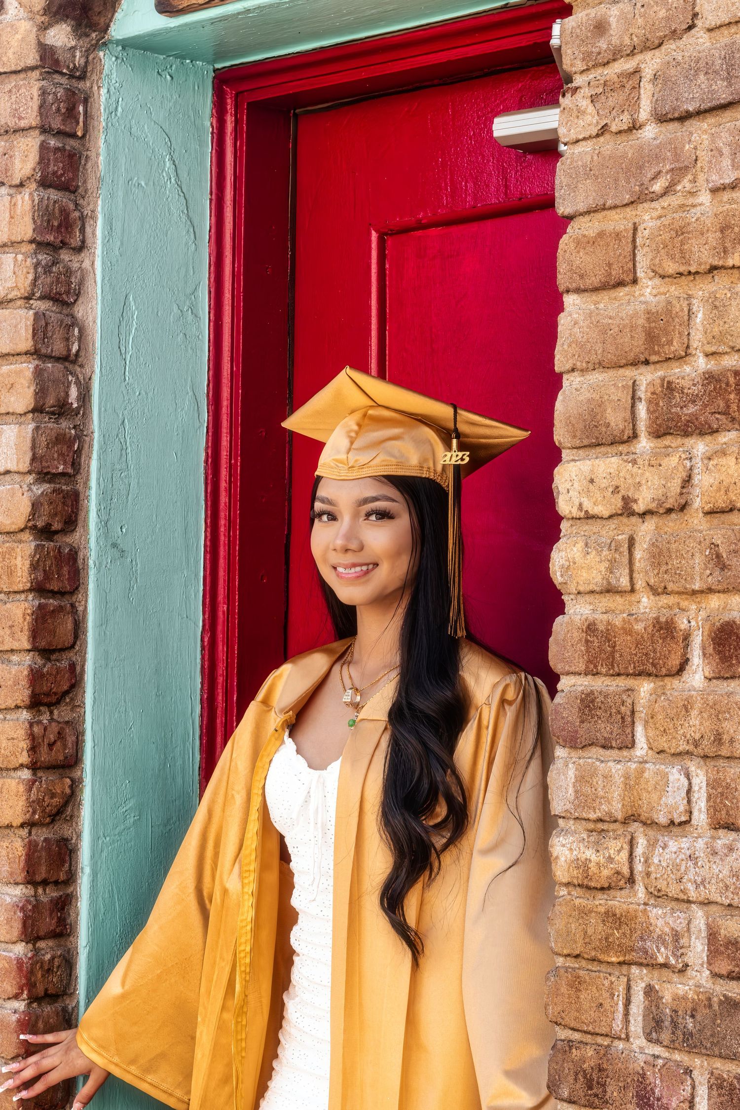 Graduate in gold cap and gown standing by red door and brick wall wearing white dress underneath.