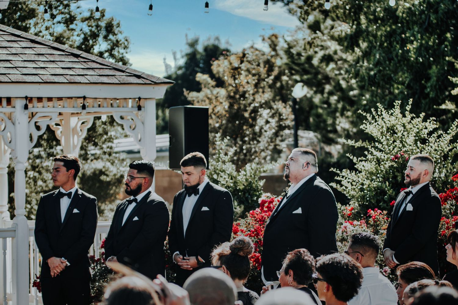 Wedding party members stand at attention in formal black suits beside a white gazebo during an outdoor ceremony.