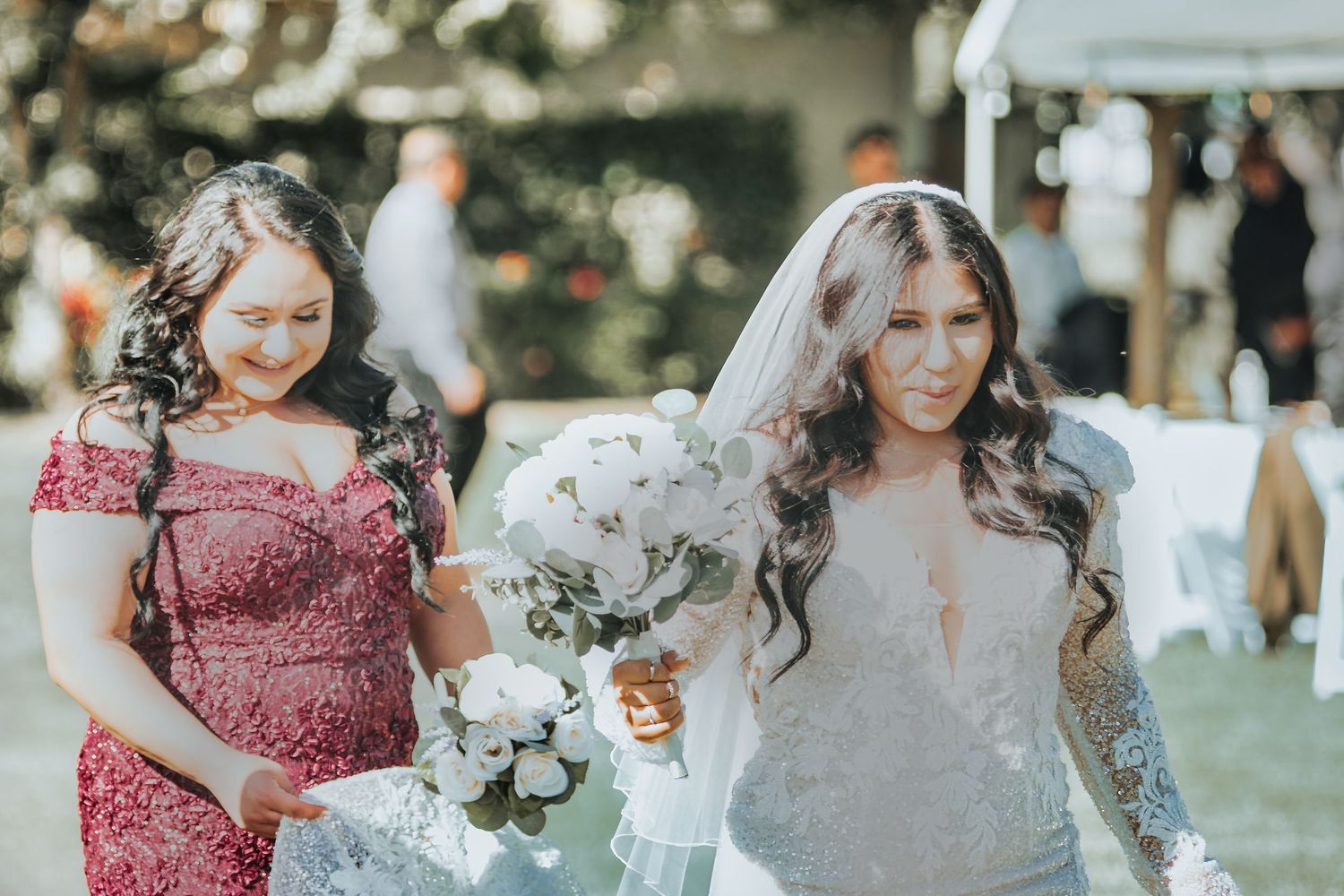 An outdoor wedding celebration shows two women walking together holding floral bouquets in a garden setting.