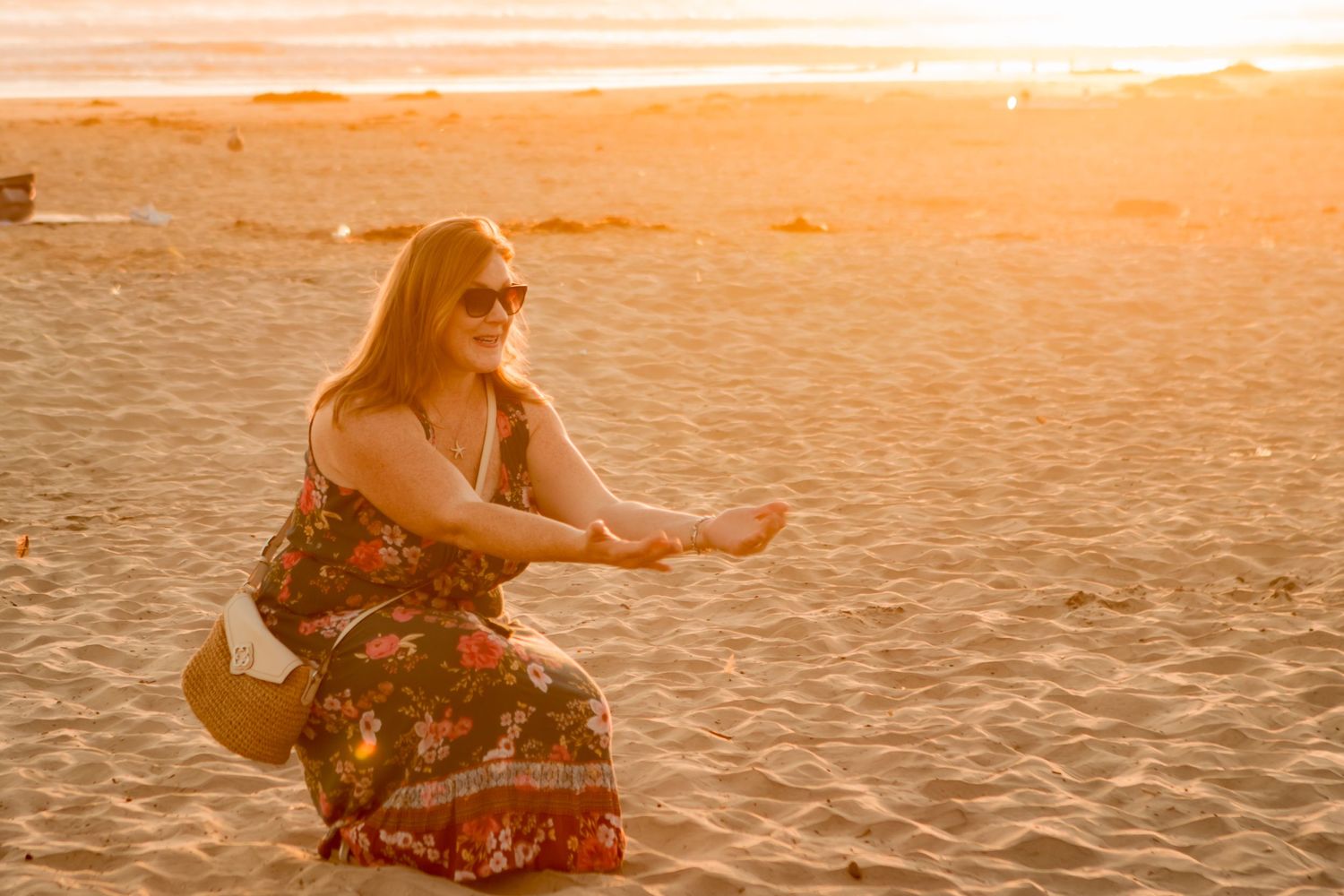 Woman in floral dress taking photos on sandy beach at sunset.