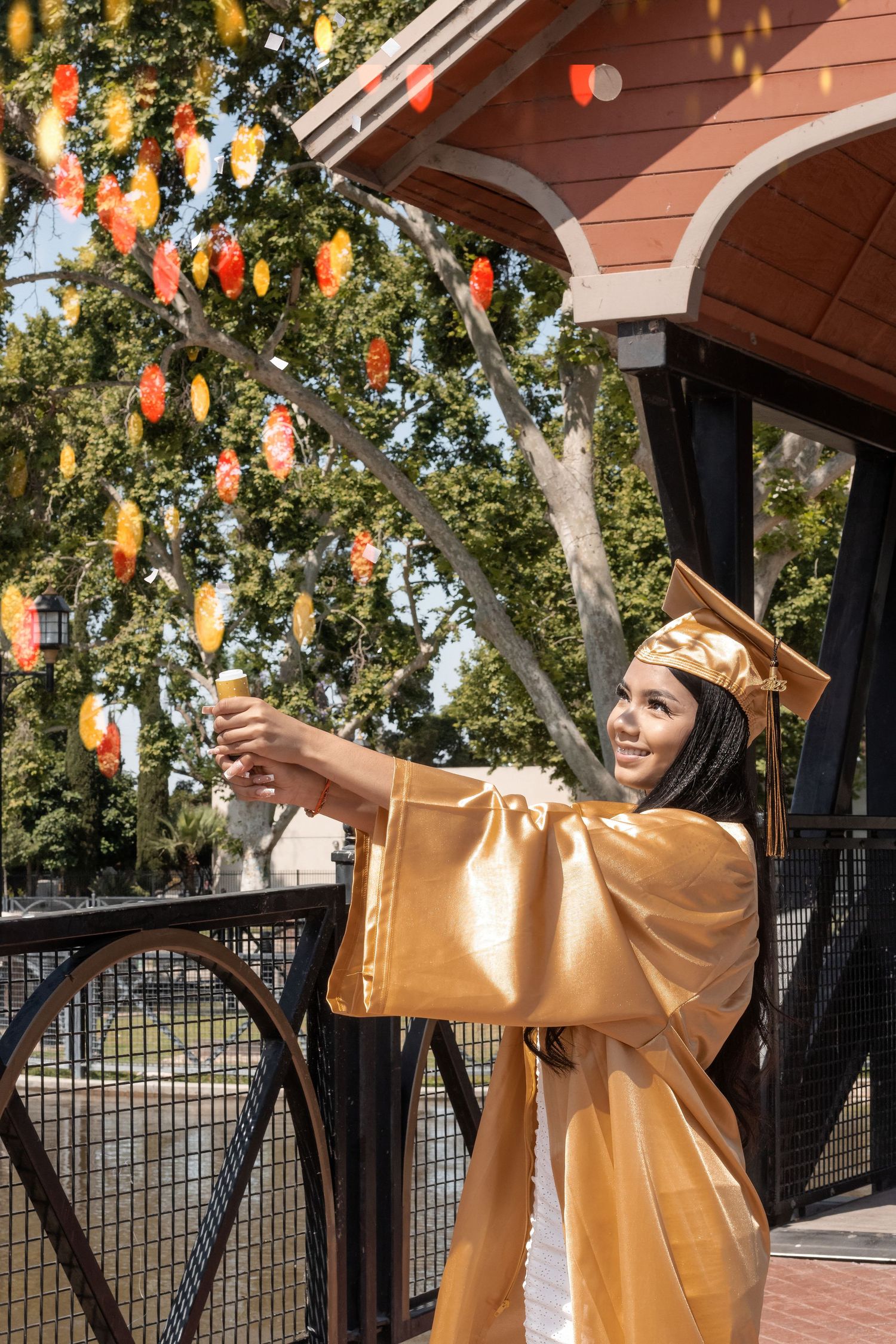 Graduate in gold cap and gown reaches upward beneath tree decorated with glowing orange lanterns during celebration.