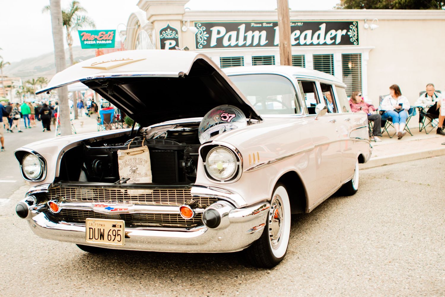 A classic 1957 Chevrolet Bel Air with its hood open on display at a vintage car show in front of Palm Traders.