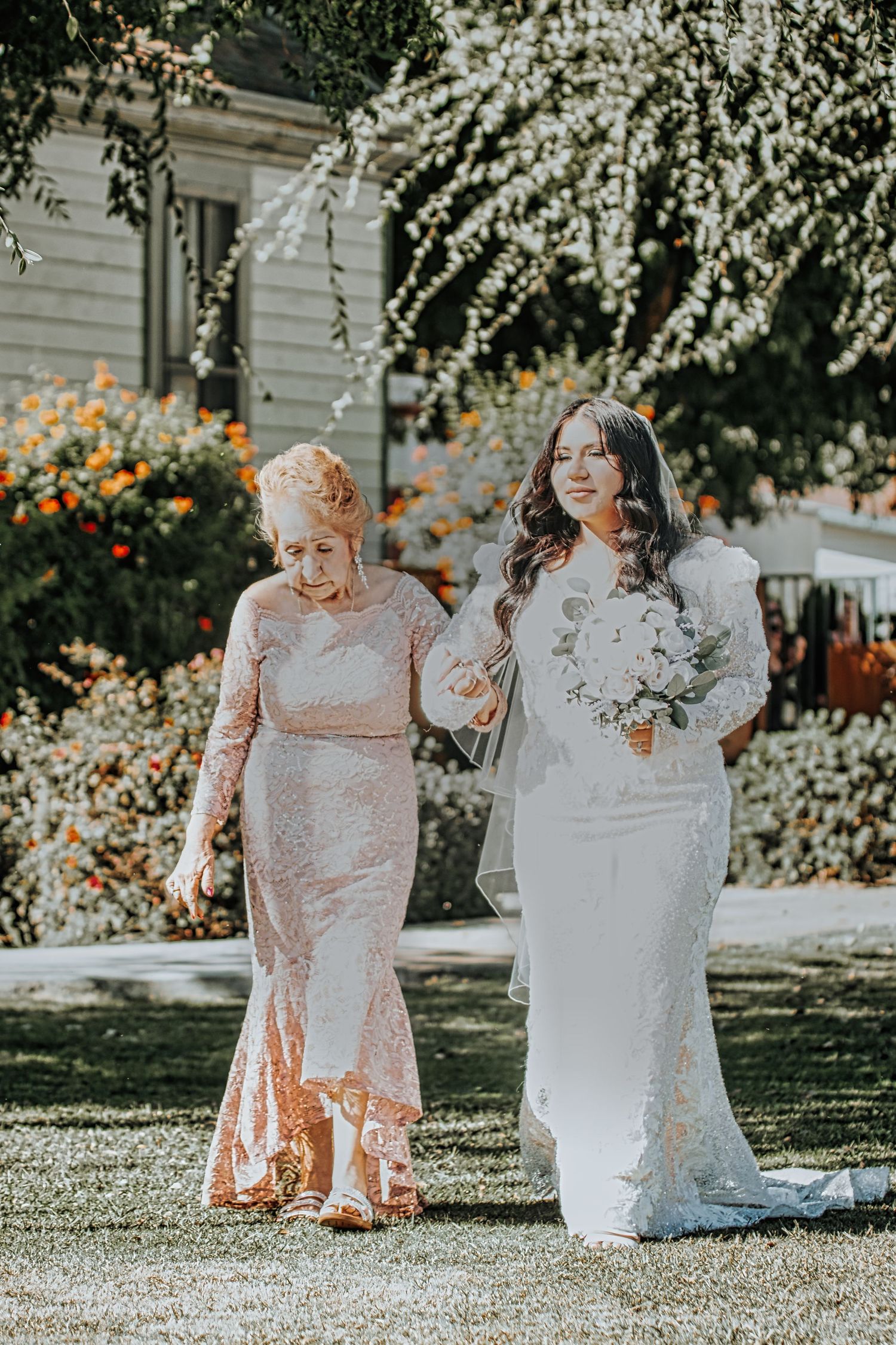 Two women in elegant lace gowns walk together in a garden setting with blooming flowers on a sunny day.