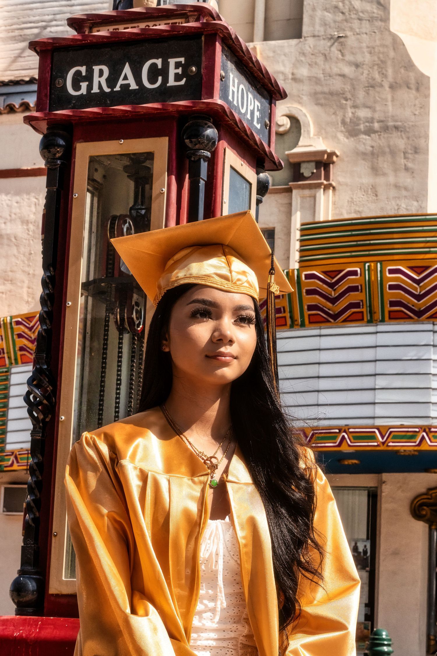 A graduate in gold cap and gown poses in front of a vintage red Grace Hotel sign with decorative pillows in the background.