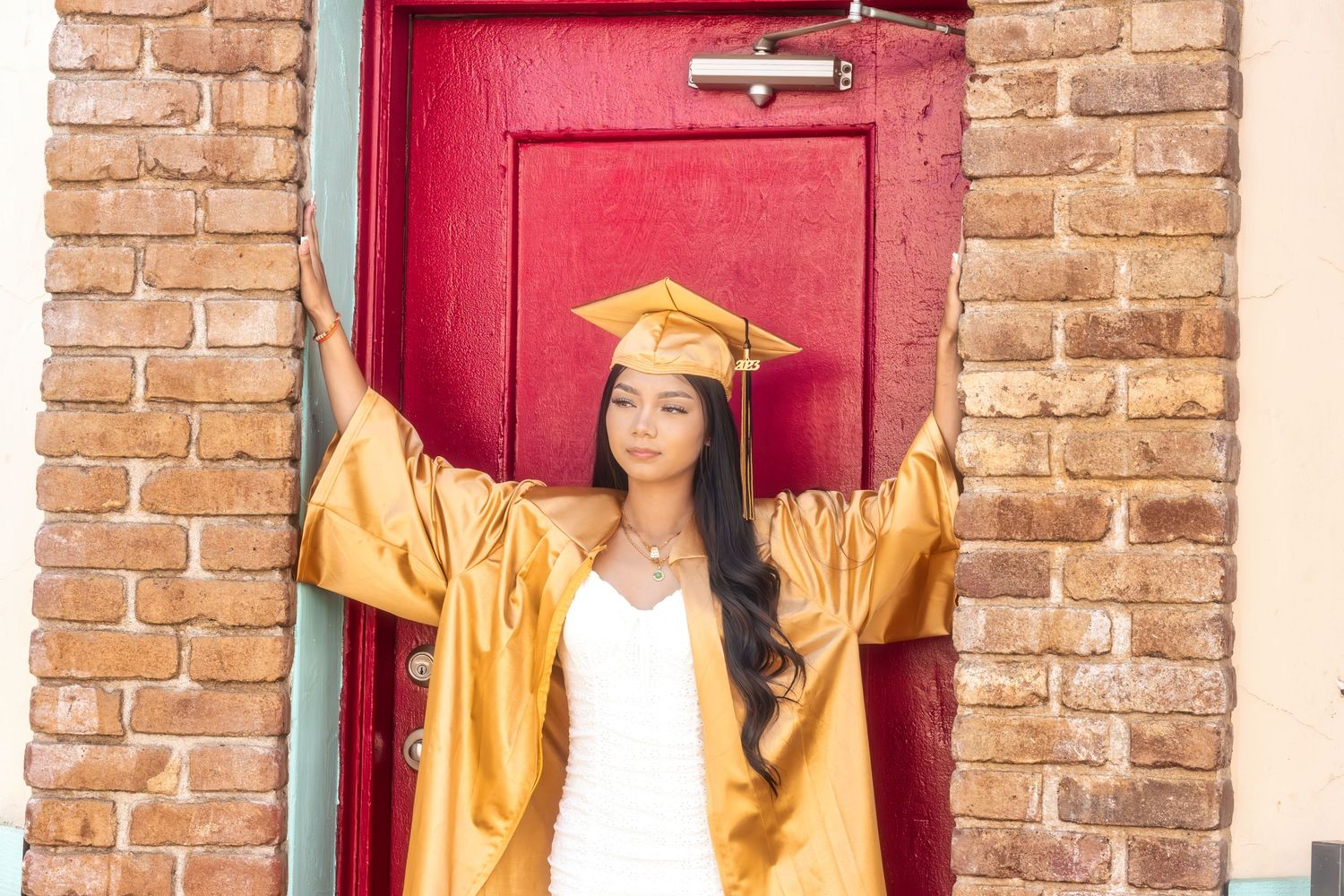A graduate in gold cap and gown poses against a red door with exposed brick wall background.