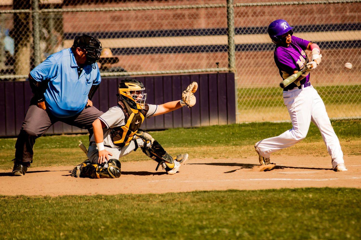 Baseball batter in purple jersey swings at pitch while catcher crouches behind home plate during daytime game.