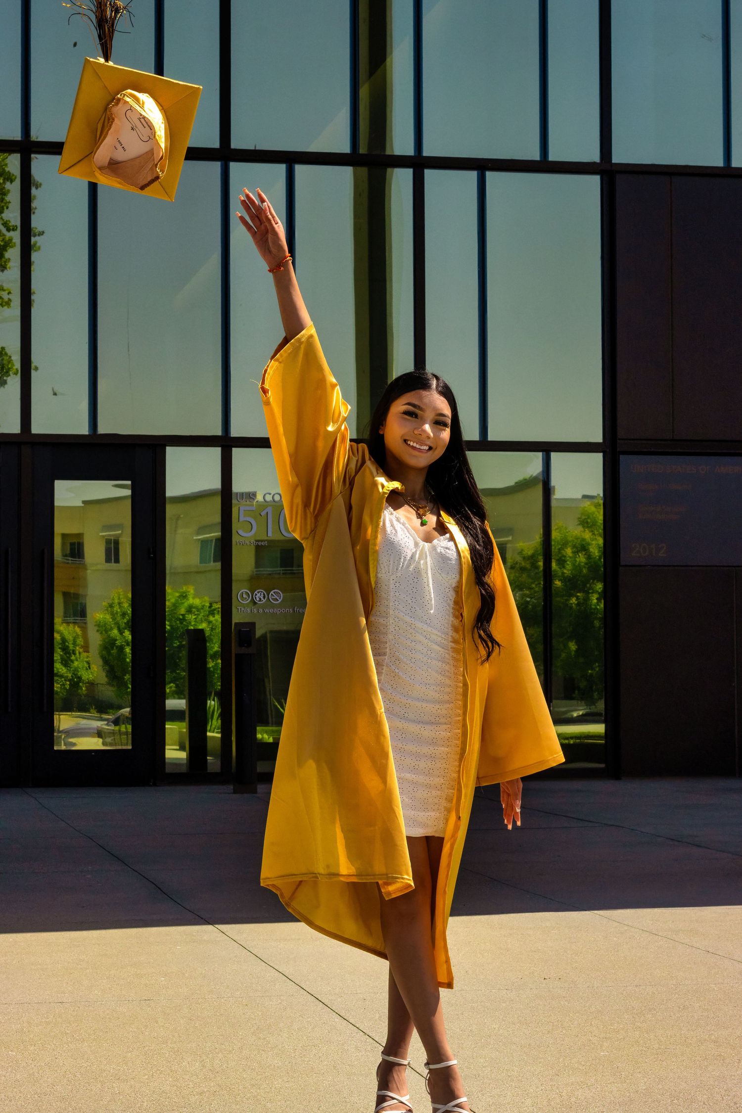 Graduate in yellow cap and gown throws graduation cap in celebration against modern building.