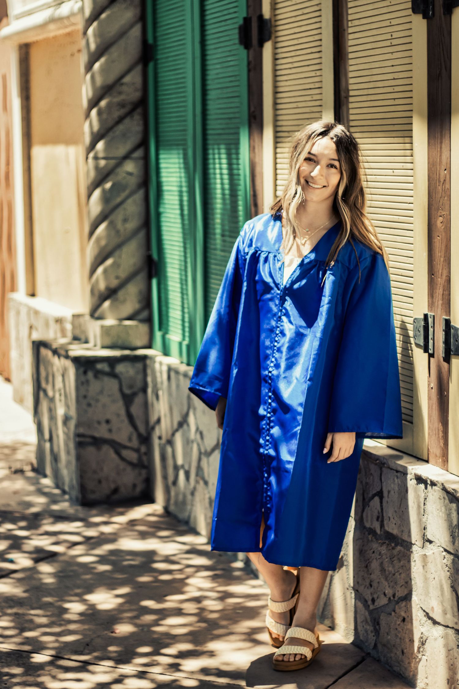 A woman stands in a bright blue satin robe and slippers against a wall with green shutters in sunlit alley.