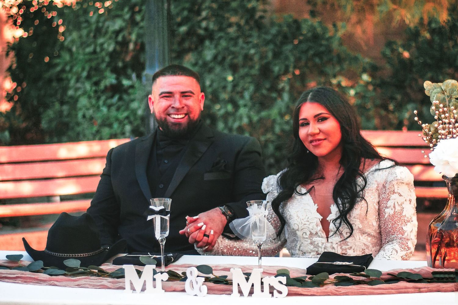 A couple sits at their wedding reception table with 'Mr & Mrs' sign displayed in front during evening festivities.