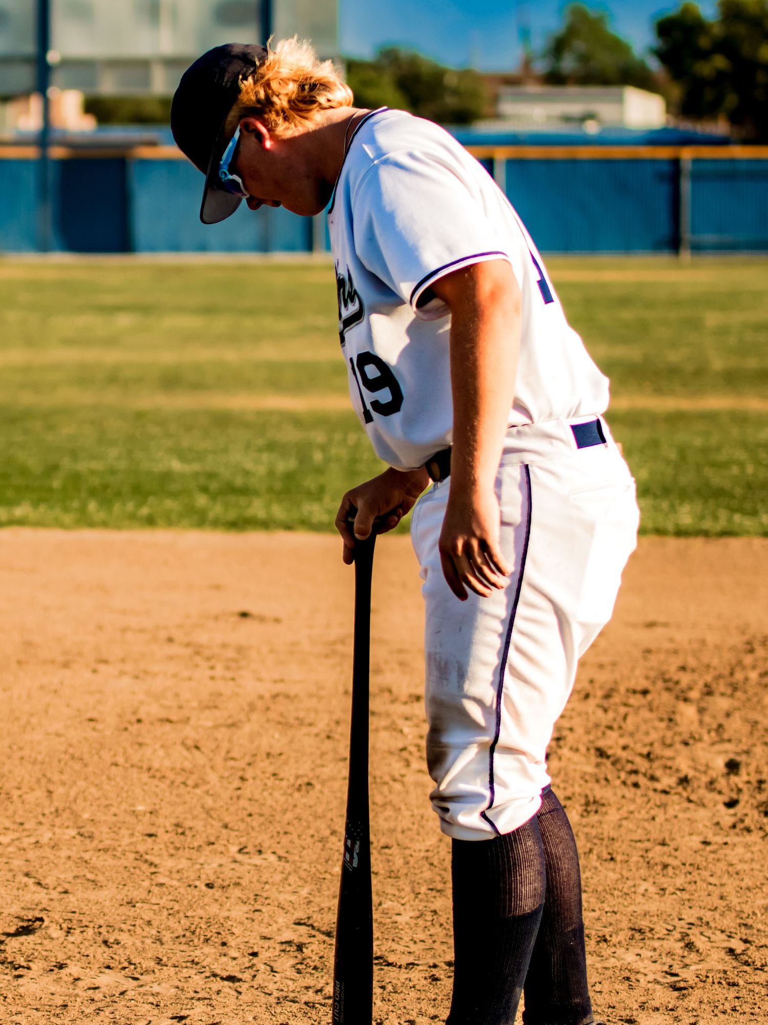 Baseball player in white uniform leans on bat during game on sunny dirt infield.