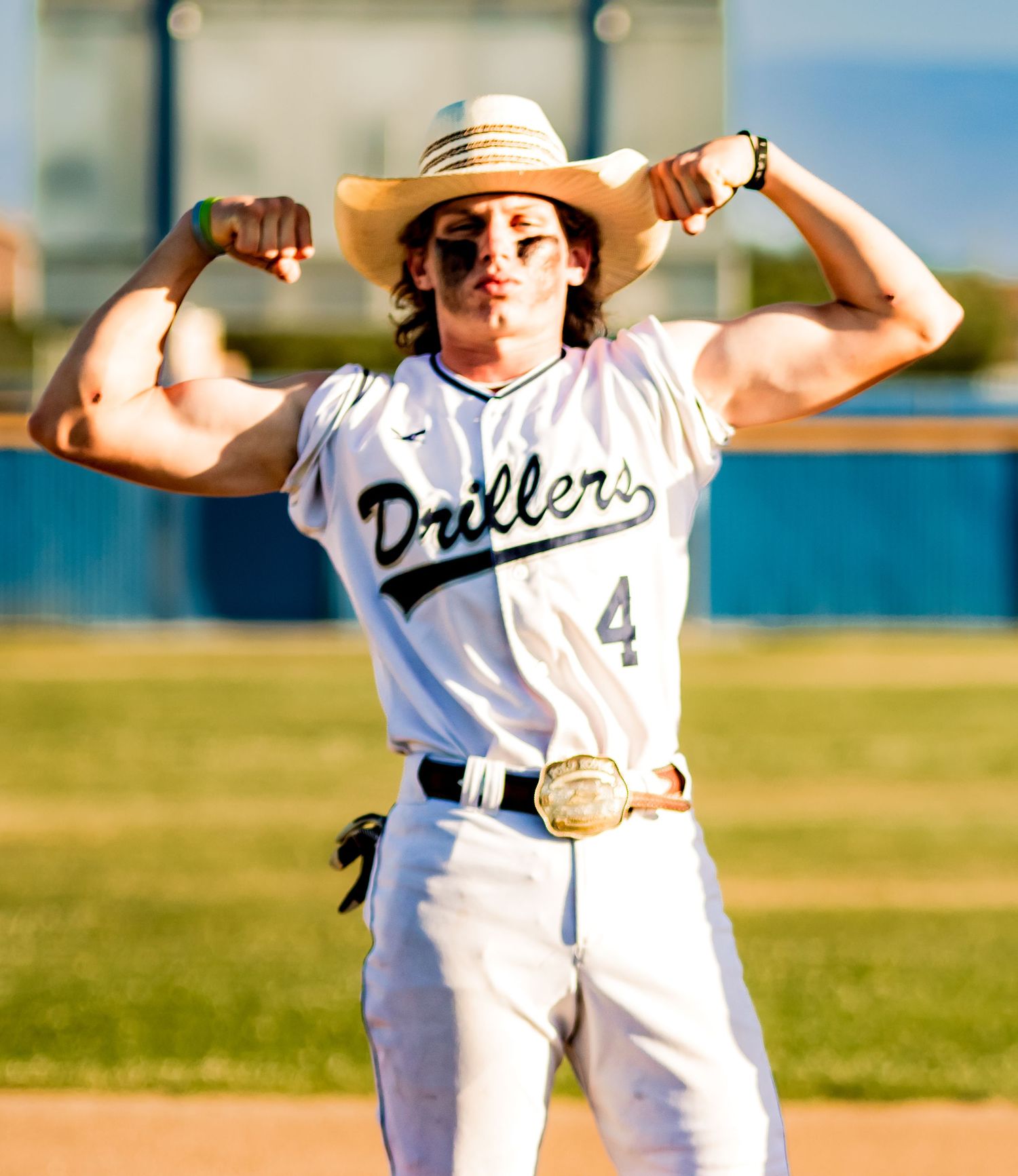 Baseball player in white Drillers uniform #4 flexes muscles while wearing a sun hat on sunny field.