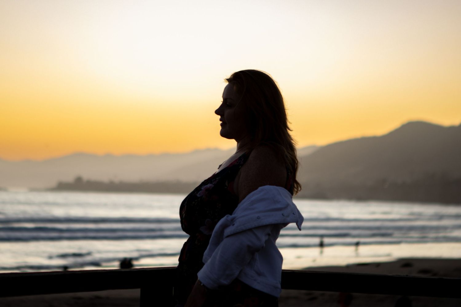 Silhouette of a person standing on beach during golden sunset with mountains in background.
