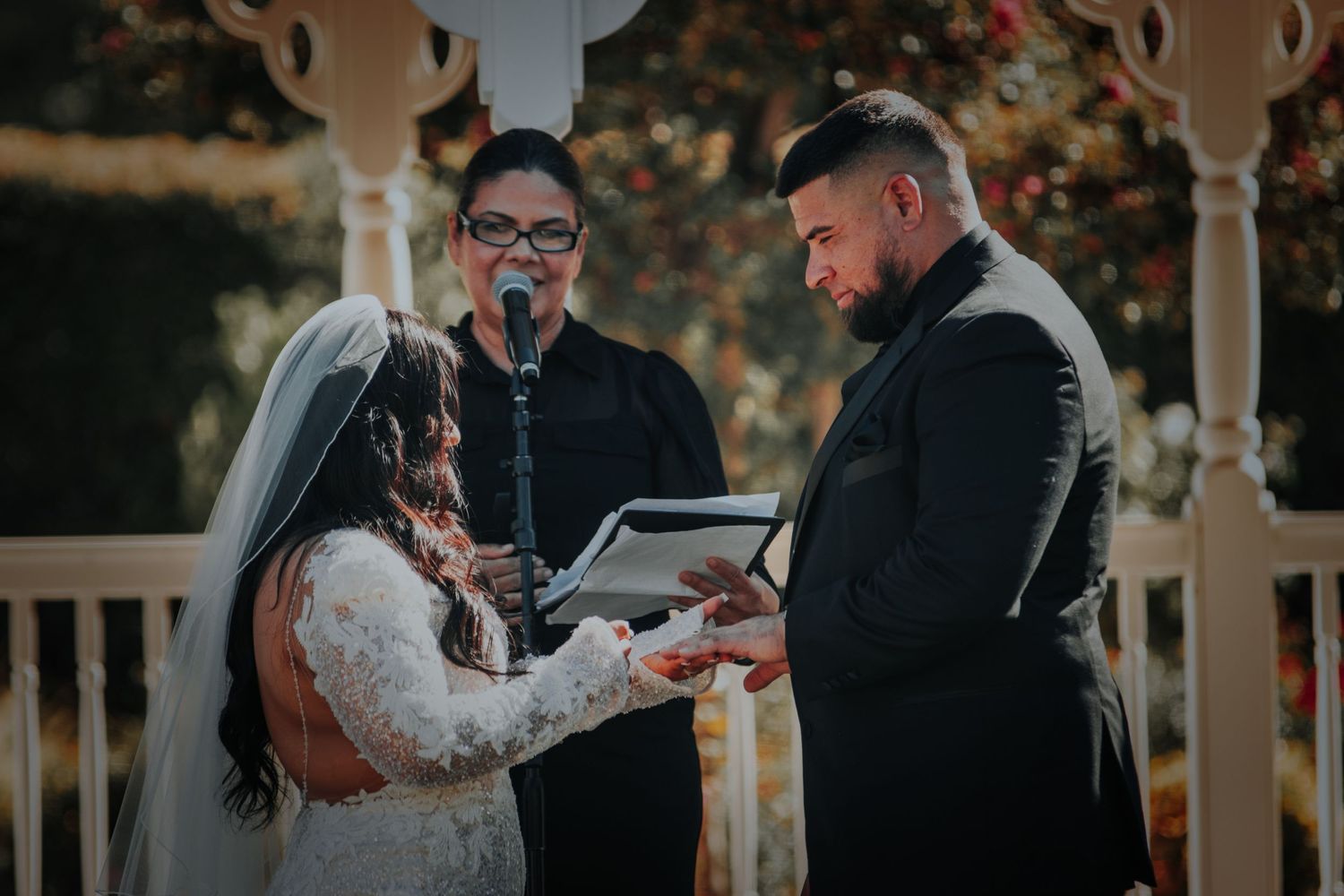 A bride and groom exchange vows during an outdoor autumn wedding ceremony under a white gazebo.