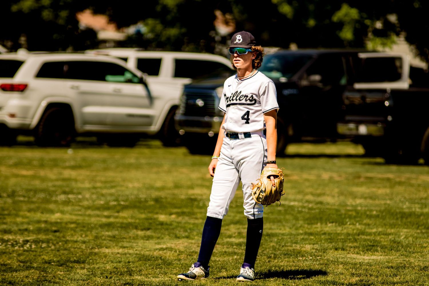 Baseball player in white uniform stands in ready position on sunny grass field with parked cars in background.