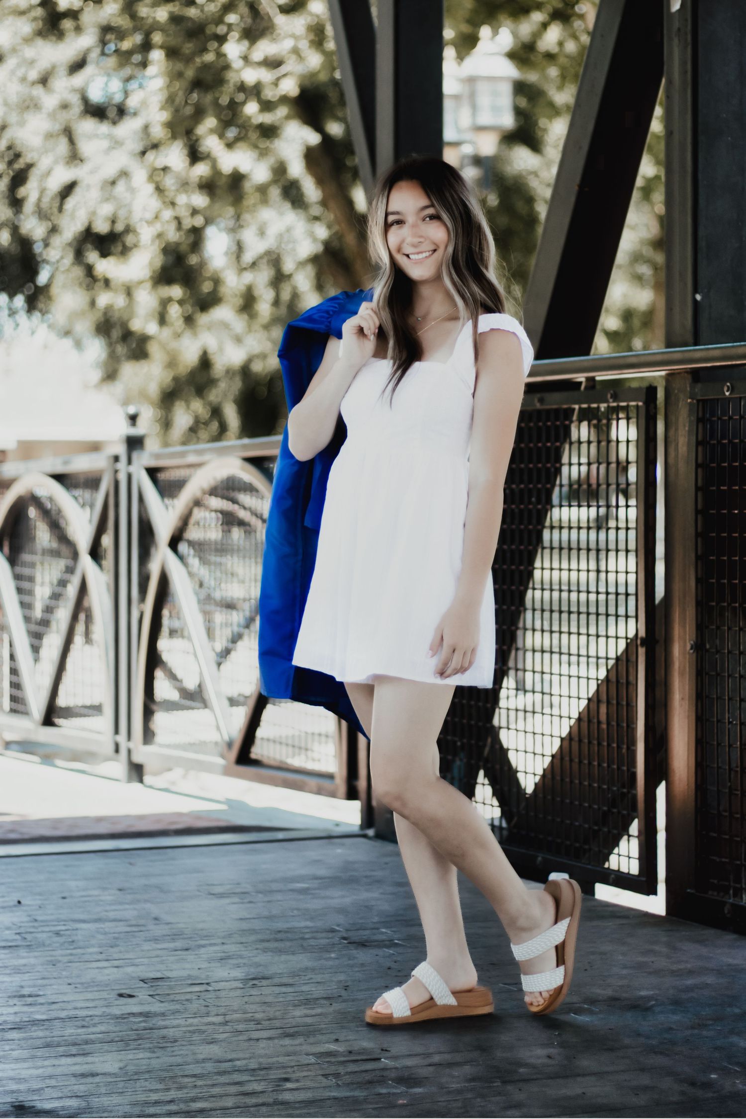A summery scene features someone in a casual white dress and sandals holding a bright blue jacket on a covered walkway.
