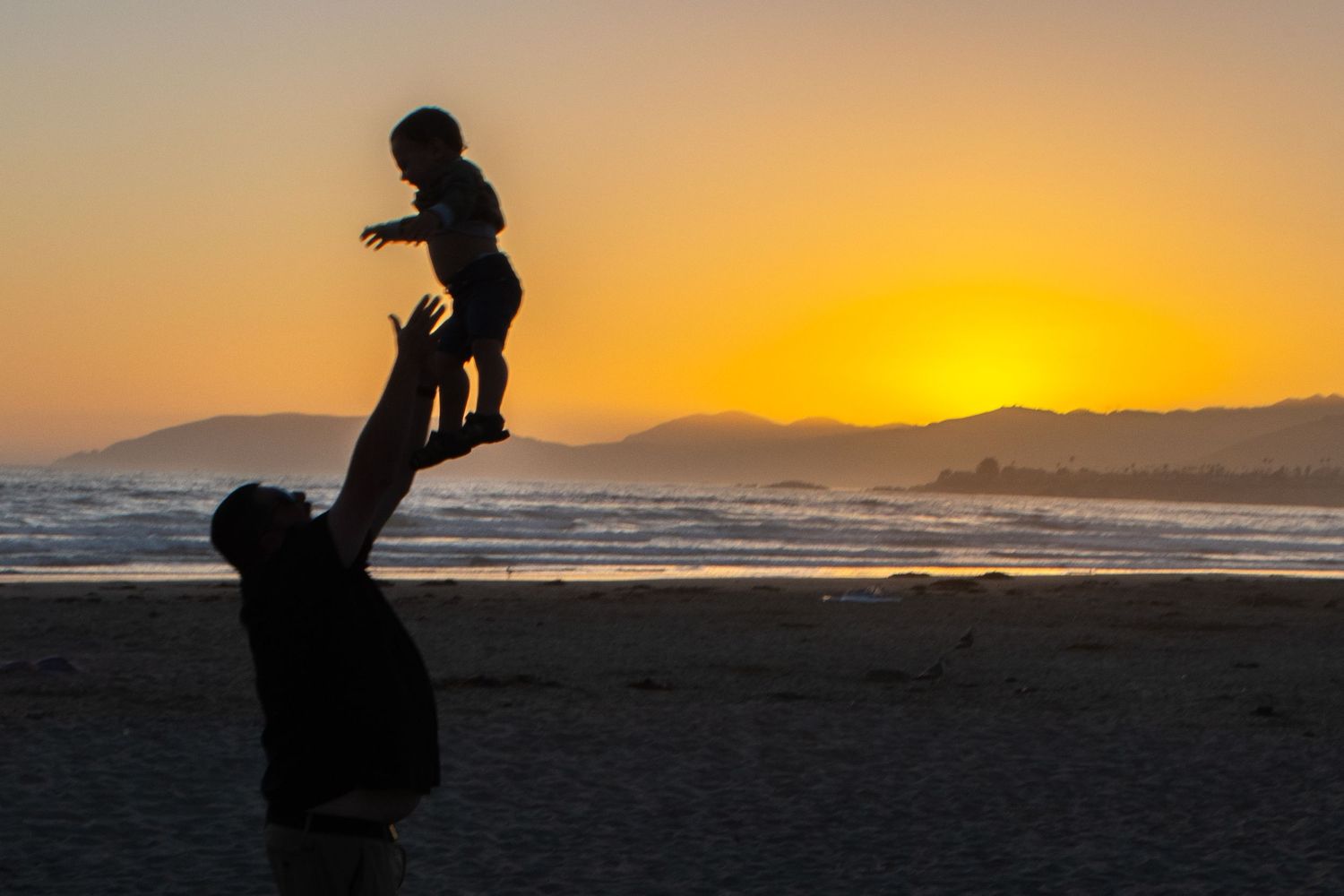 Silhouette of parent lifting child against orange sunset at beach.