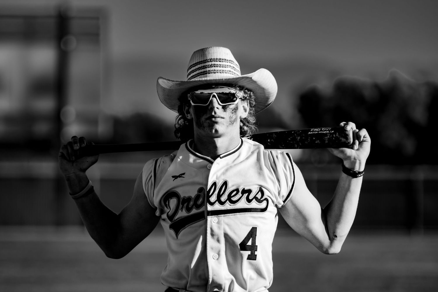 Black and white portrait of a softball player wearing sunglasses and cowboy hat holding bat across shoulders in Ballers jersey.