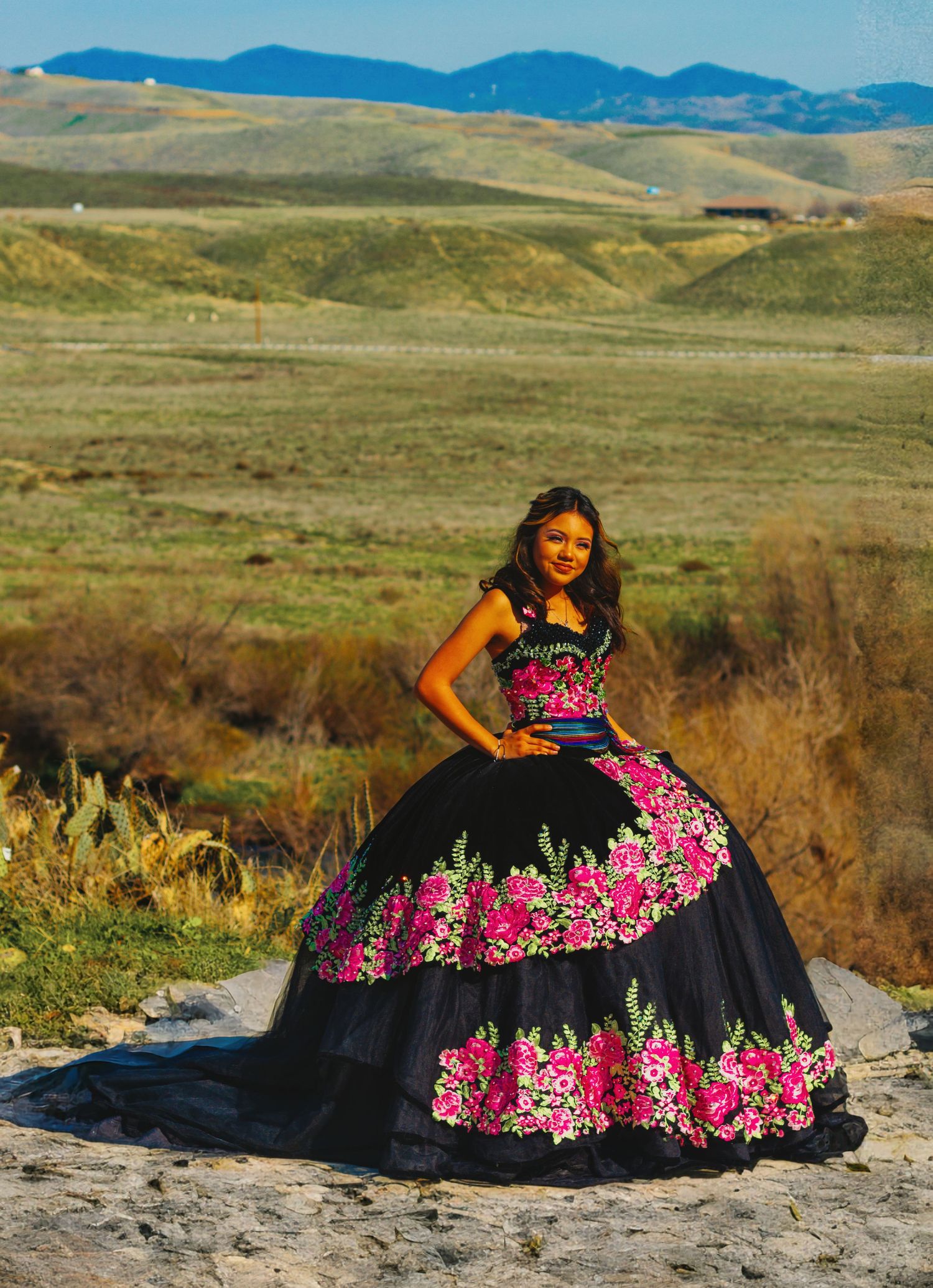 A stunning black and pink floral quinceañera dress stands out against rolling hills and mountains in the background.