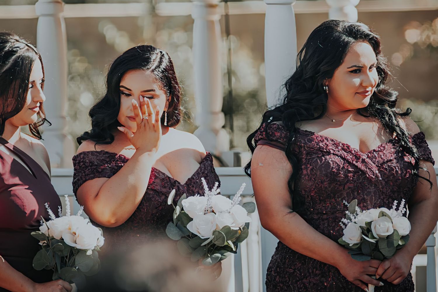 Bridesmaids in burgundy off-shoulder dresses hold white bouquets during an outdoor wedding ceremony.