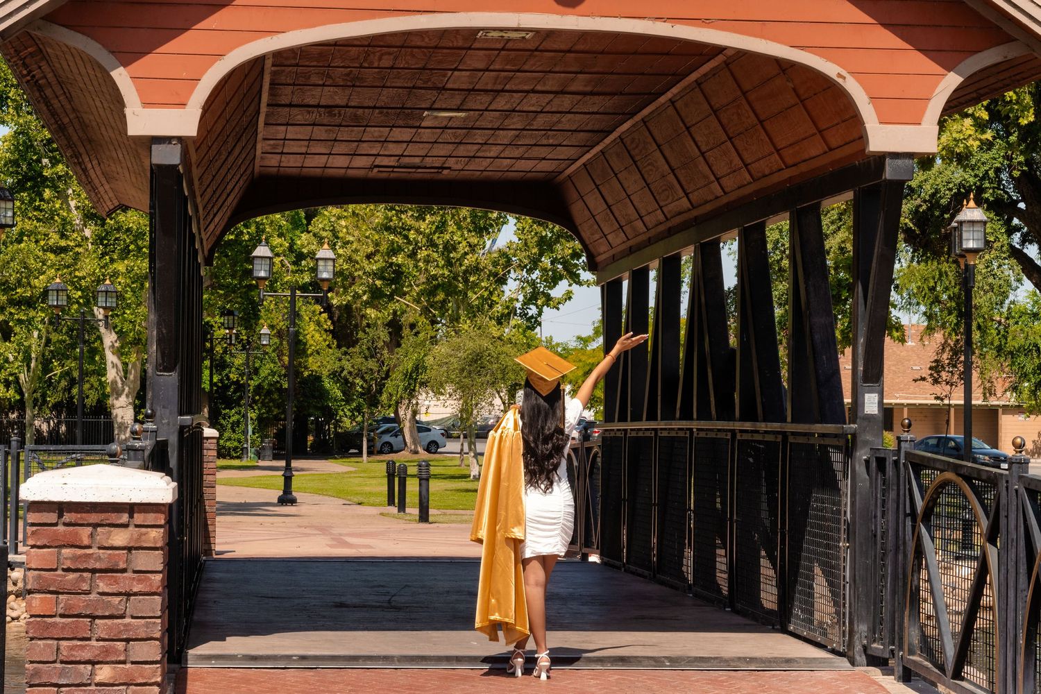 A person in a yellow dress stands under a covered bridge walkway with brick pillars and green trees on a sunny day.