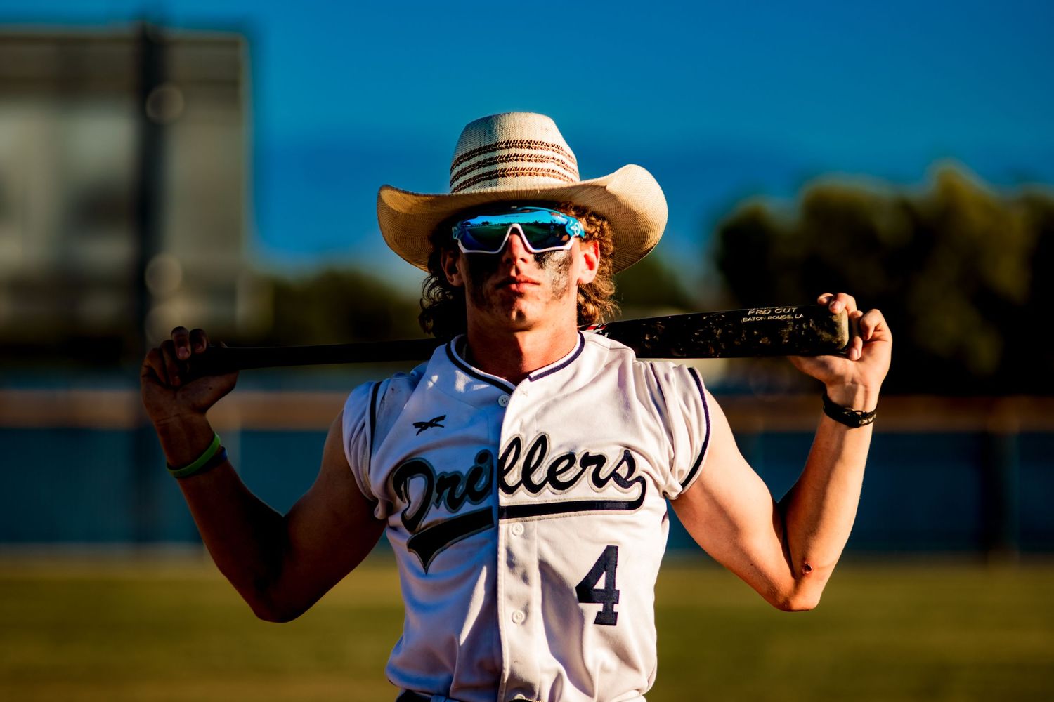 Softball player wearing white Bulldogs uniform and cowboy hat holds bat across shoulders against blue sky background.