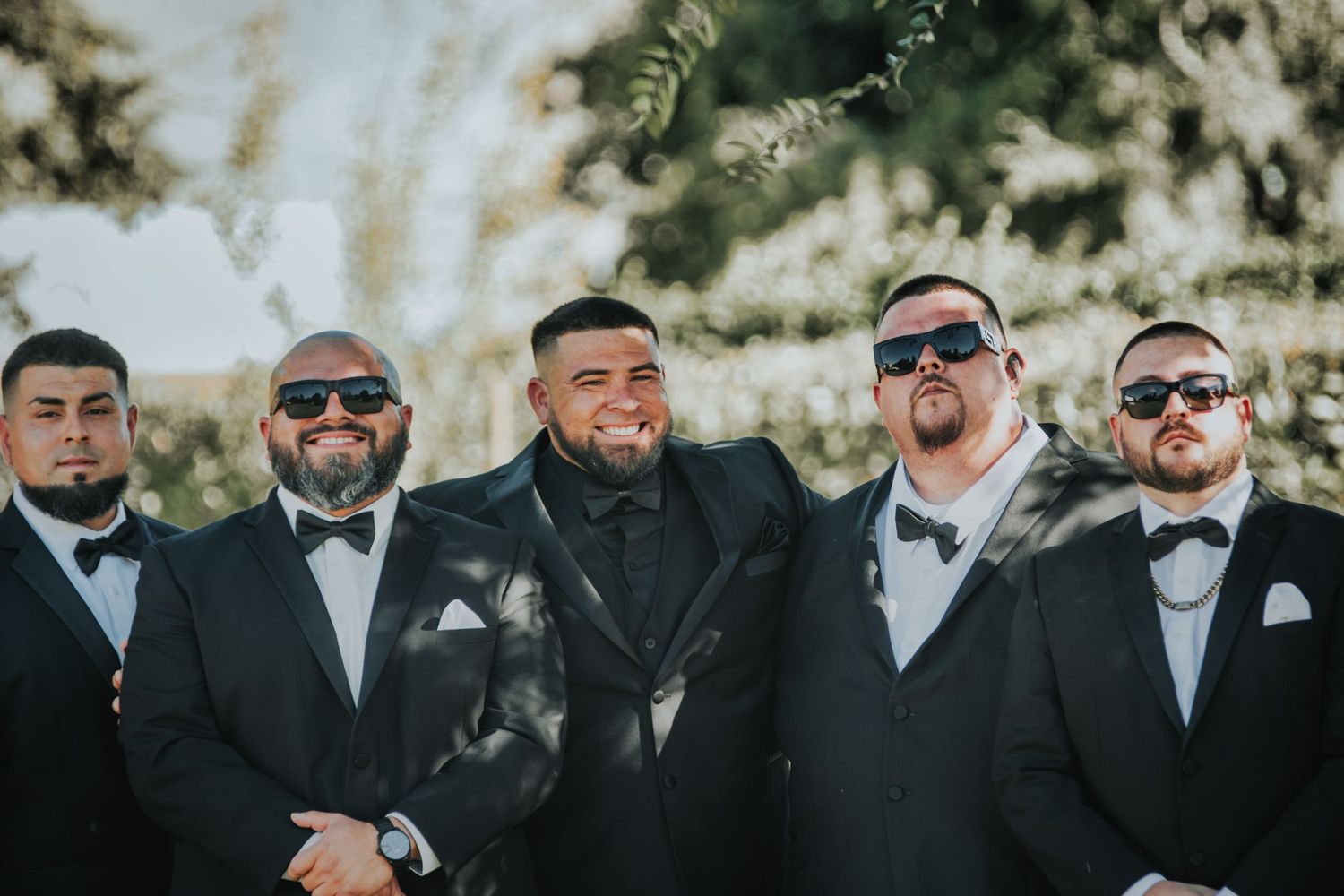 A group of groomsmen in black tuxedos and bowties laugh together during an outdoor wedding photo session.