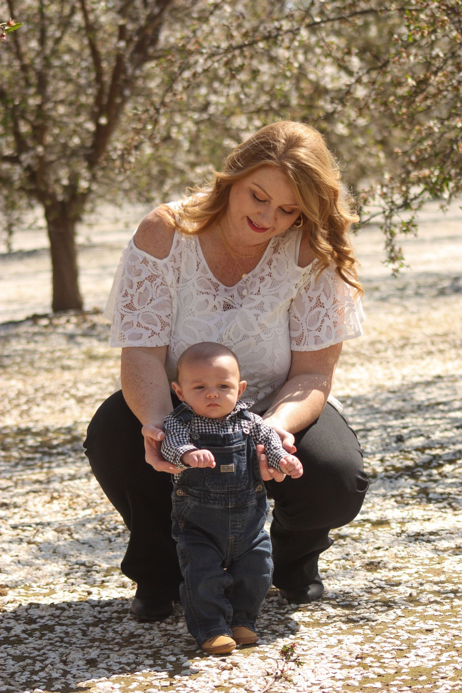 A white lace-dressed woman holding a baby in denim overalls among blooming almond trees.