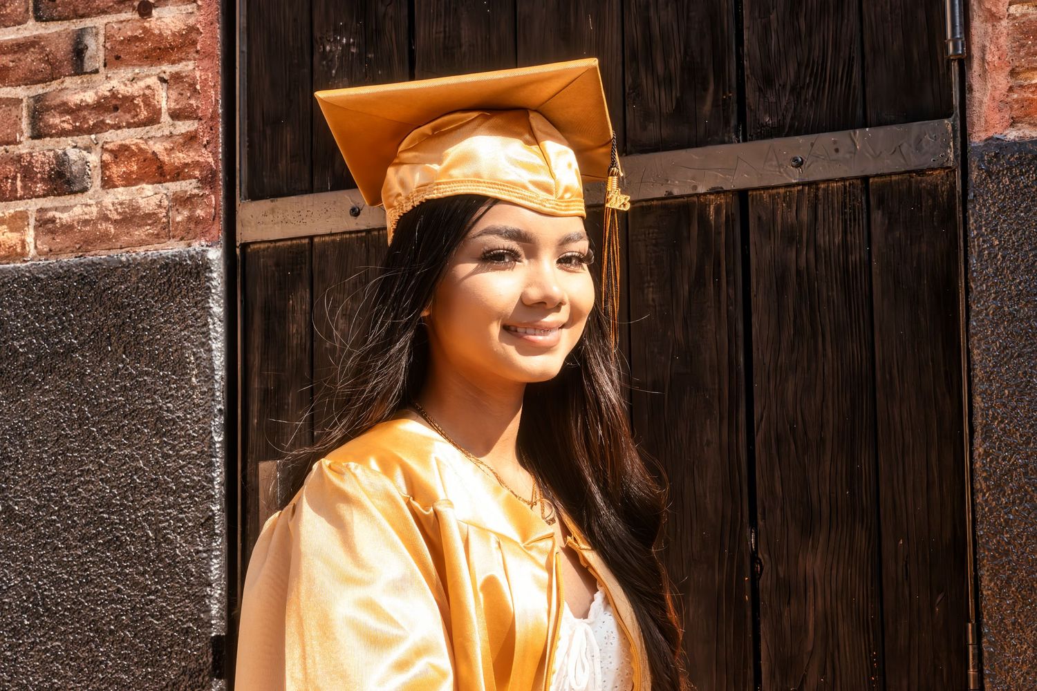 Graduate wearing gold cap and gown stands smiling in front of rustic brick and wooden wall.