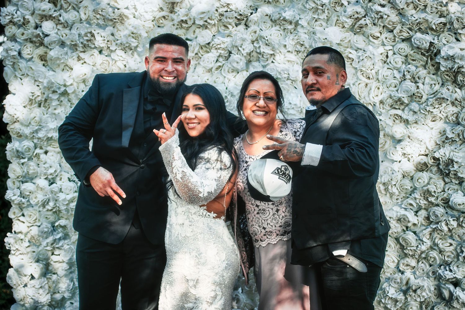 A happy wedding celebration with a couple and two guests against a white floral backdrop.