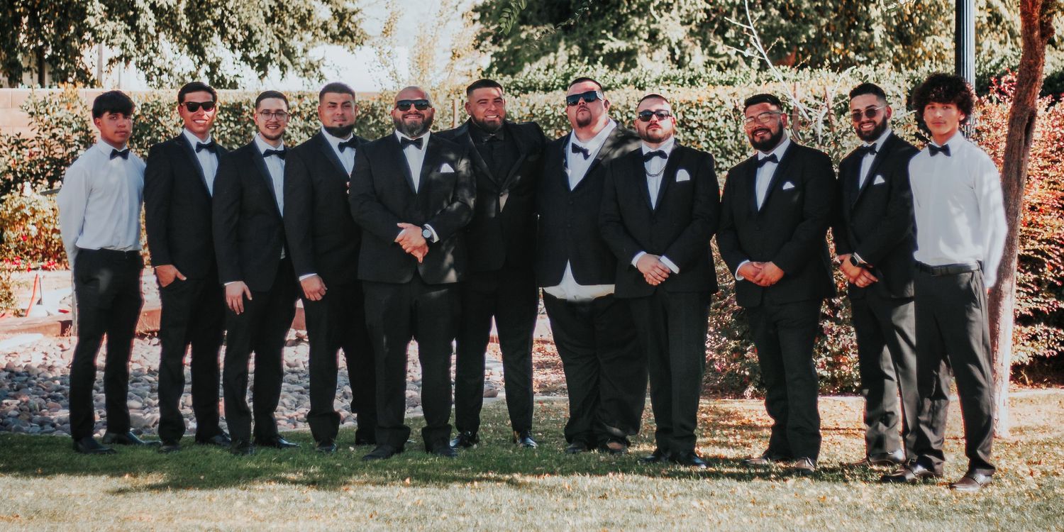 A large groomsmen party in black suits and tuxedos posing together outdoors.