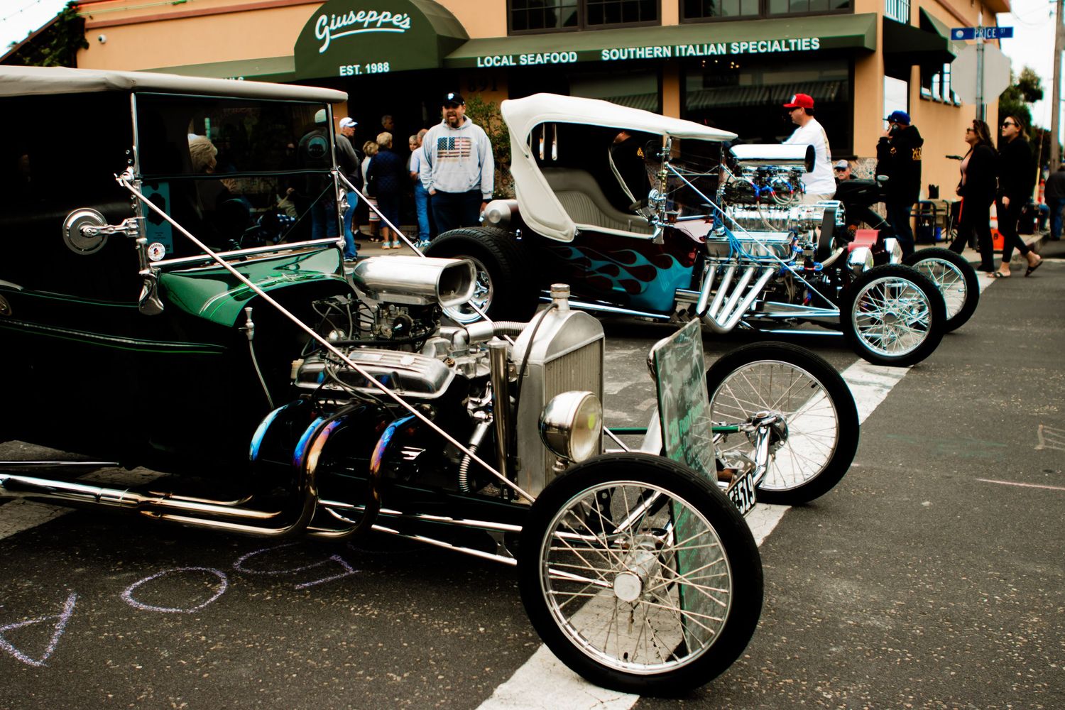 Custom hot rods and vintage cars lined up at a car show outside a restaurant with a green awning.