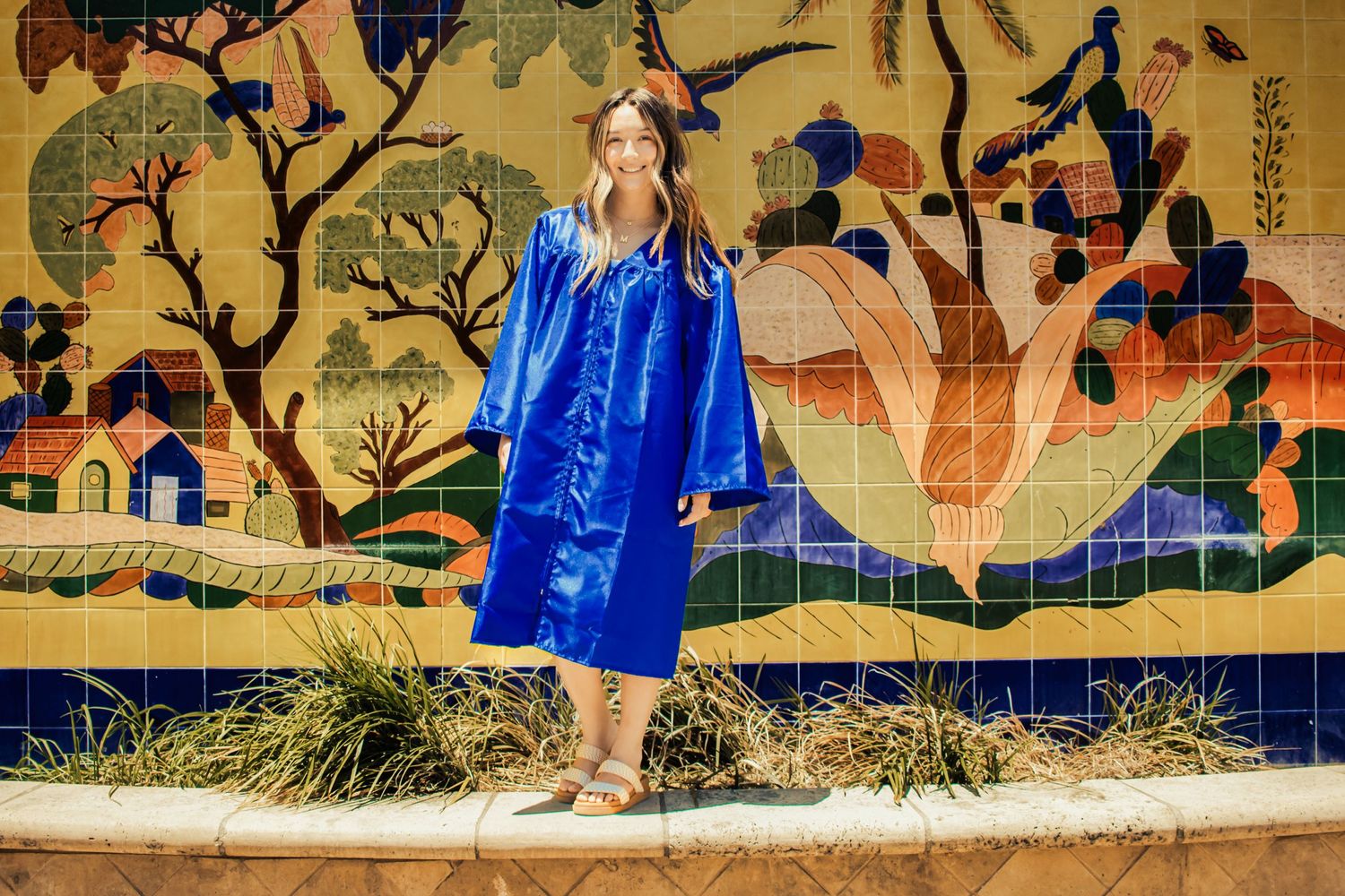 A bright blue graduation gown stands out against a colorful wall mural featuring trees and houses in a whimsical scene.