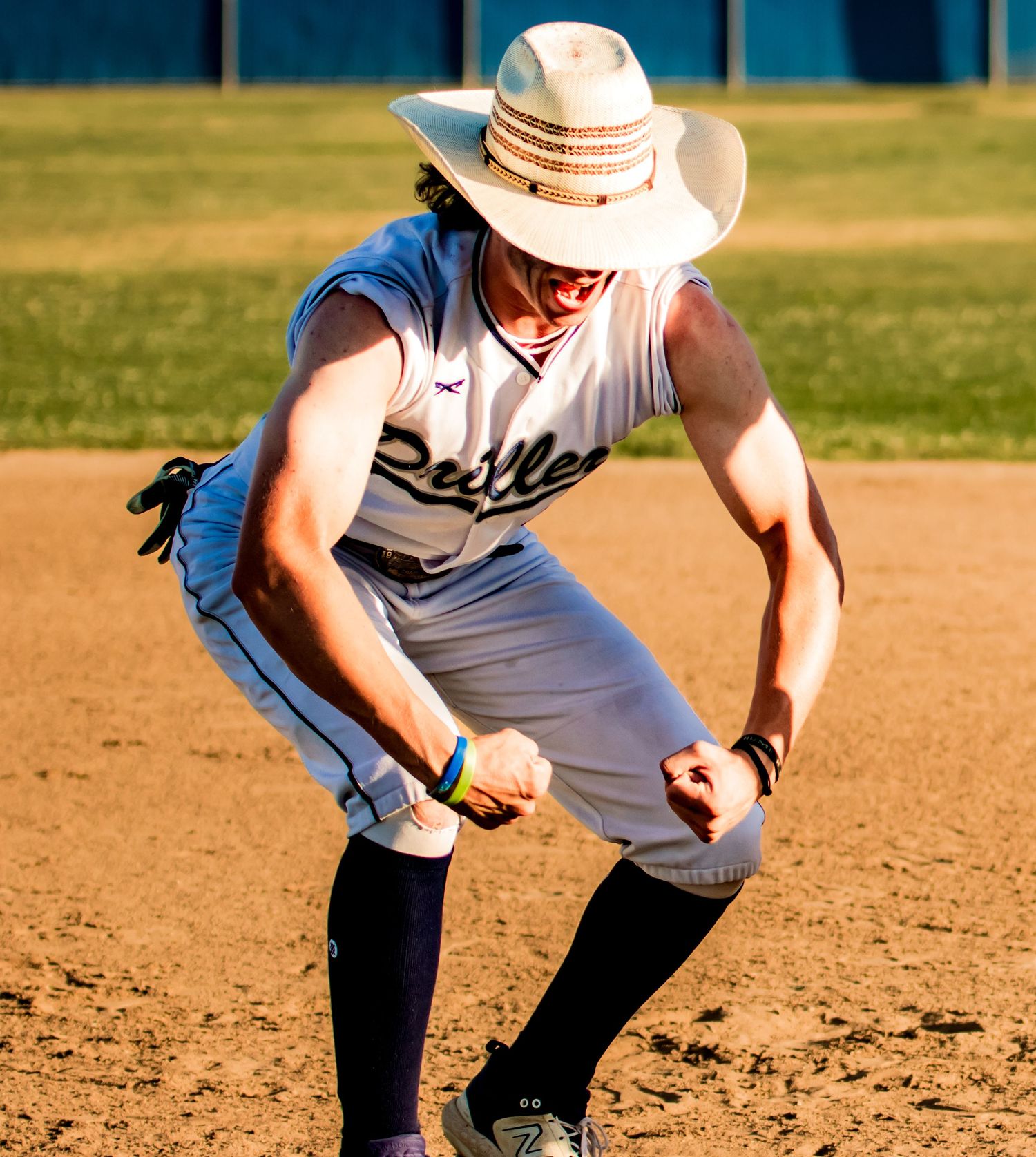Baseball player in a white uniform and cowboy hat ready in fielding position on a dirt infield during sunset.