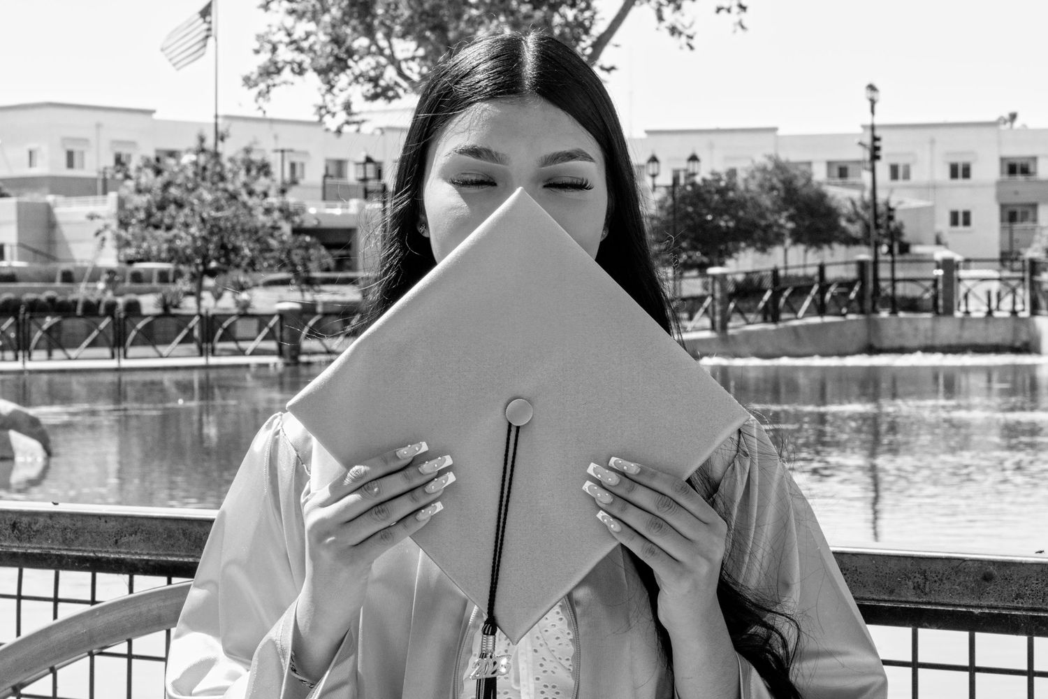 A graduate student holds up their diploma in front of their face while standing near a pool in a black and white photo.