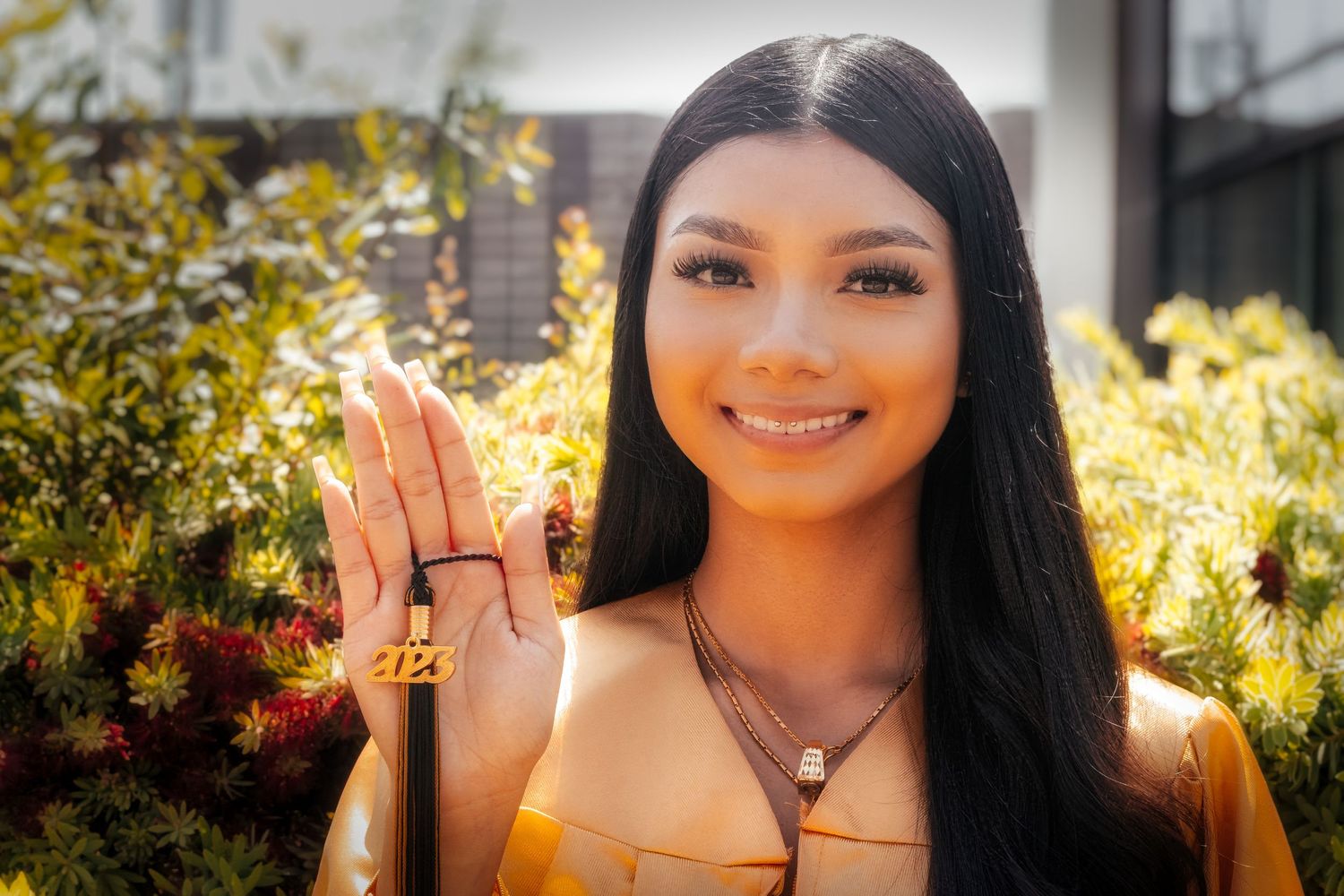 A person with long dark hair holds up their hand showing a ring while standing outdoors near yellow flowers.