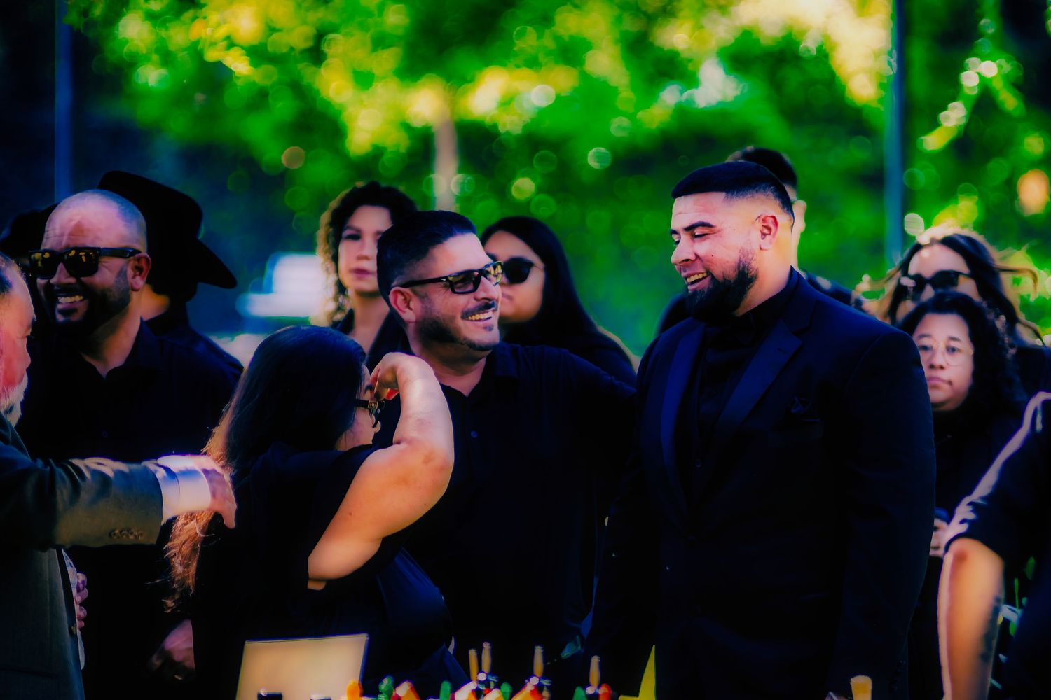A group of guests dressed in dark attire gather outdoors at an evening social event surrounded by green foliage.