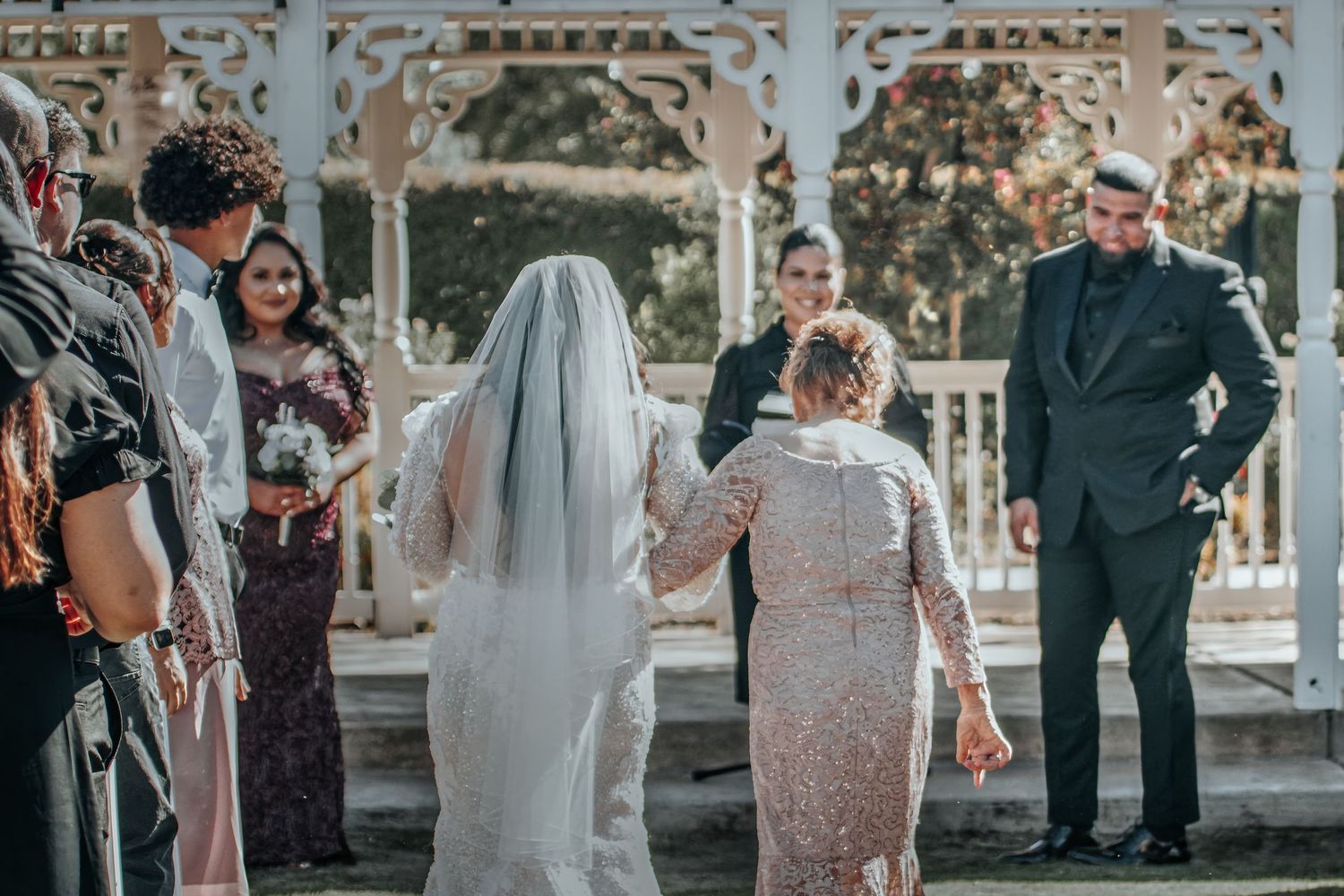 A small outdoor wedding ceremony takes place at an elegant white gazebo surrounded by family members.
