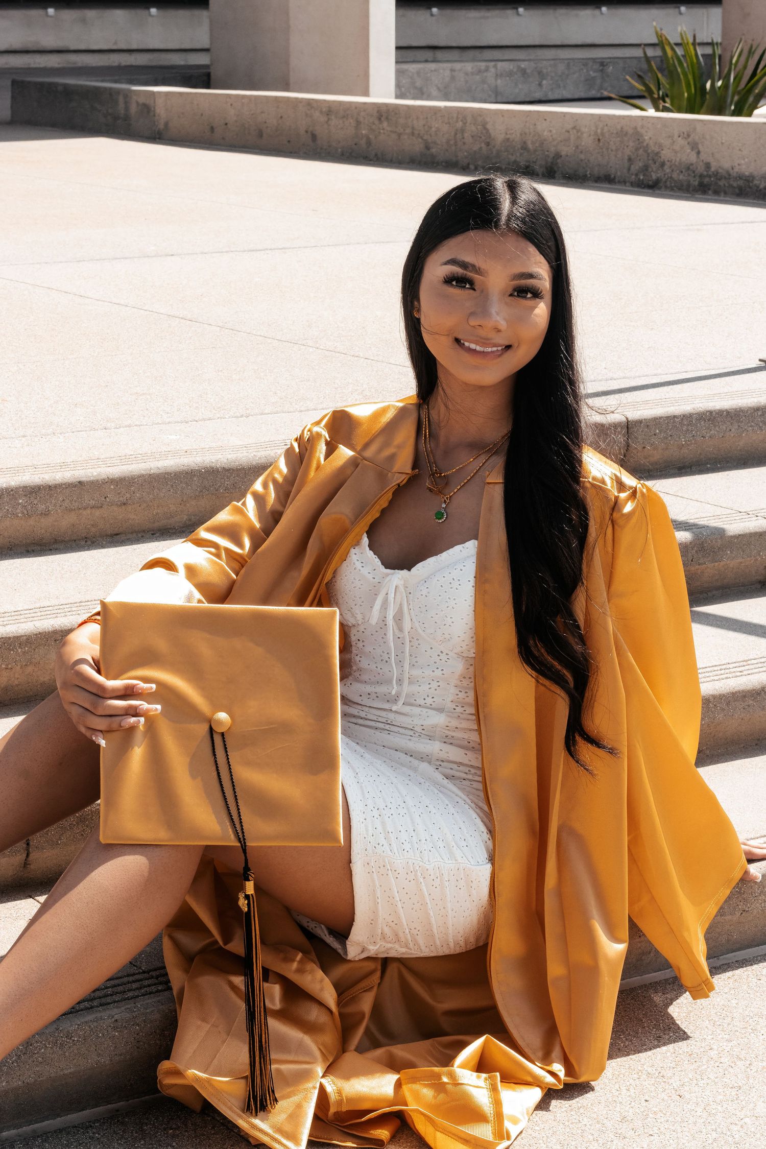 A model in a white dress poses on steps with a yellow coat and matching handbag in warm sunlit setting.