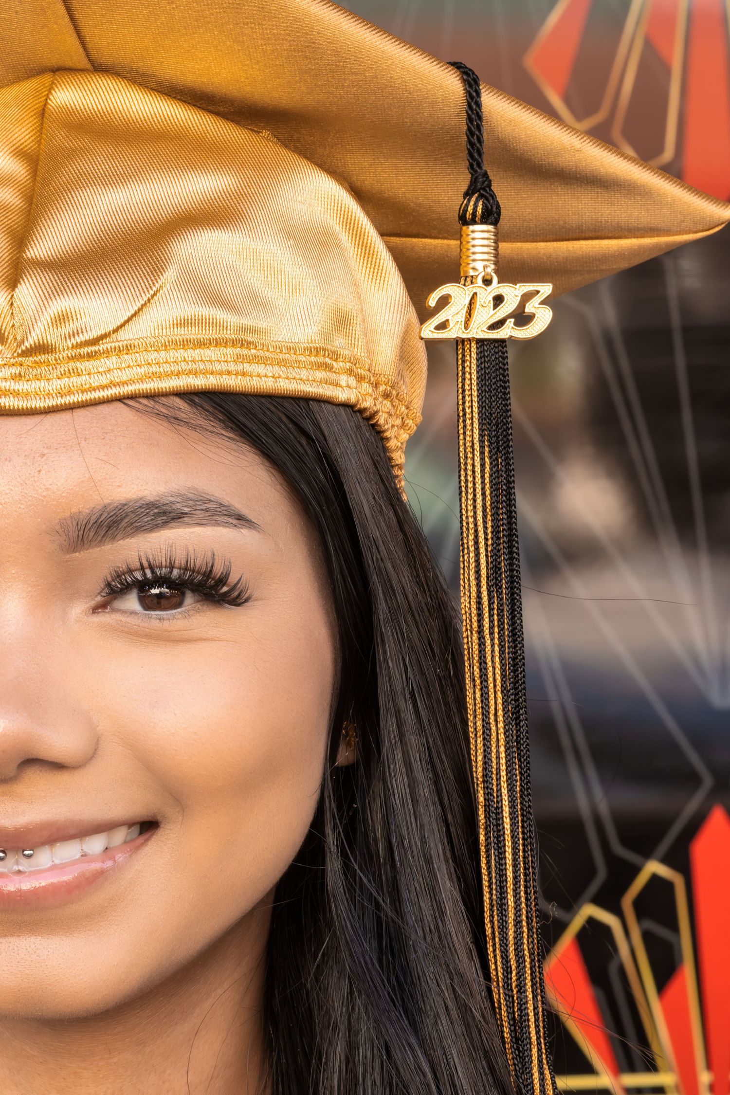 Graduate wearing gold cap and tassel with class of 2023 charm against decorative background.