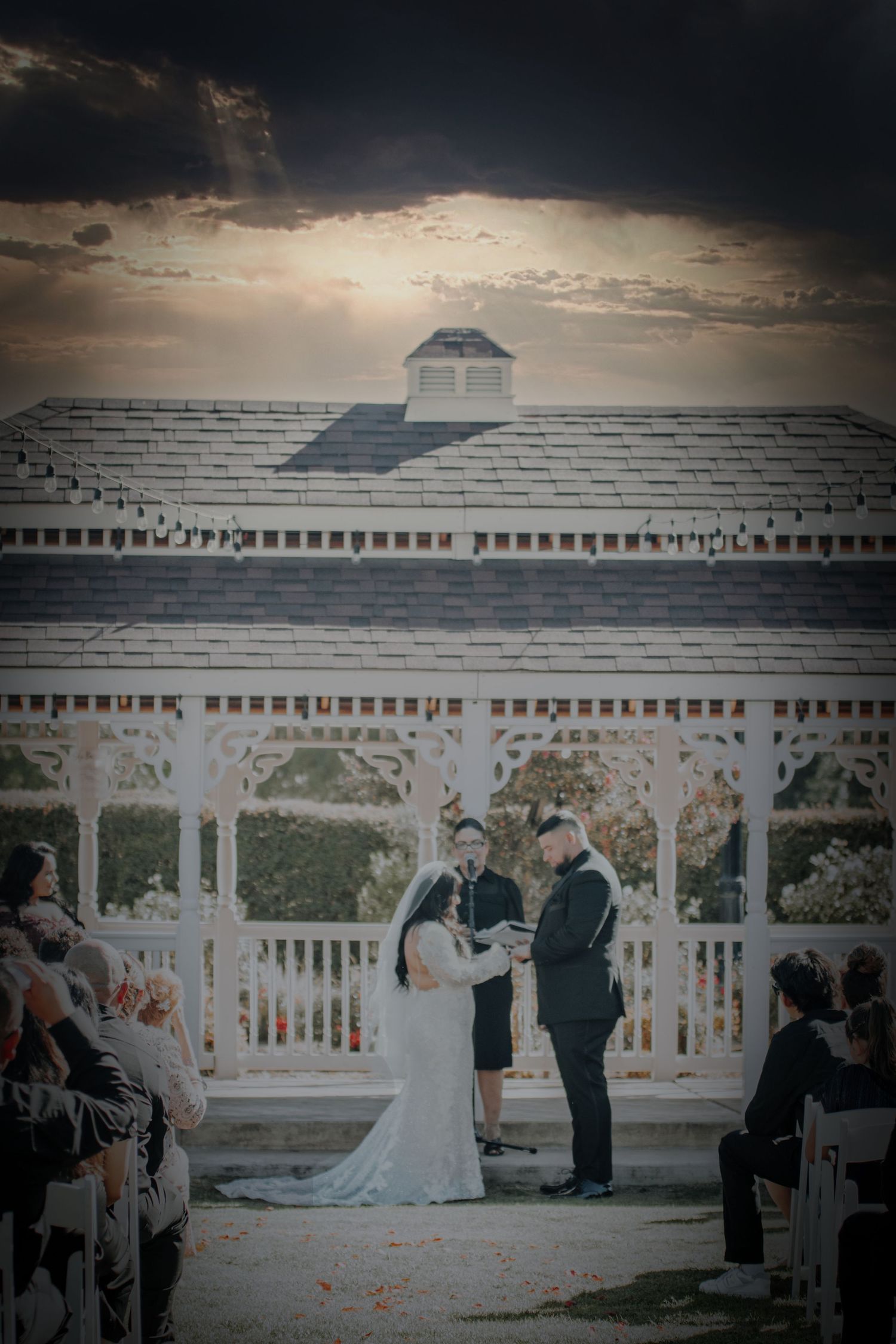 A wedding ceremony takes place at a white gazebo with dramatic storm clouds overhead at sunset.